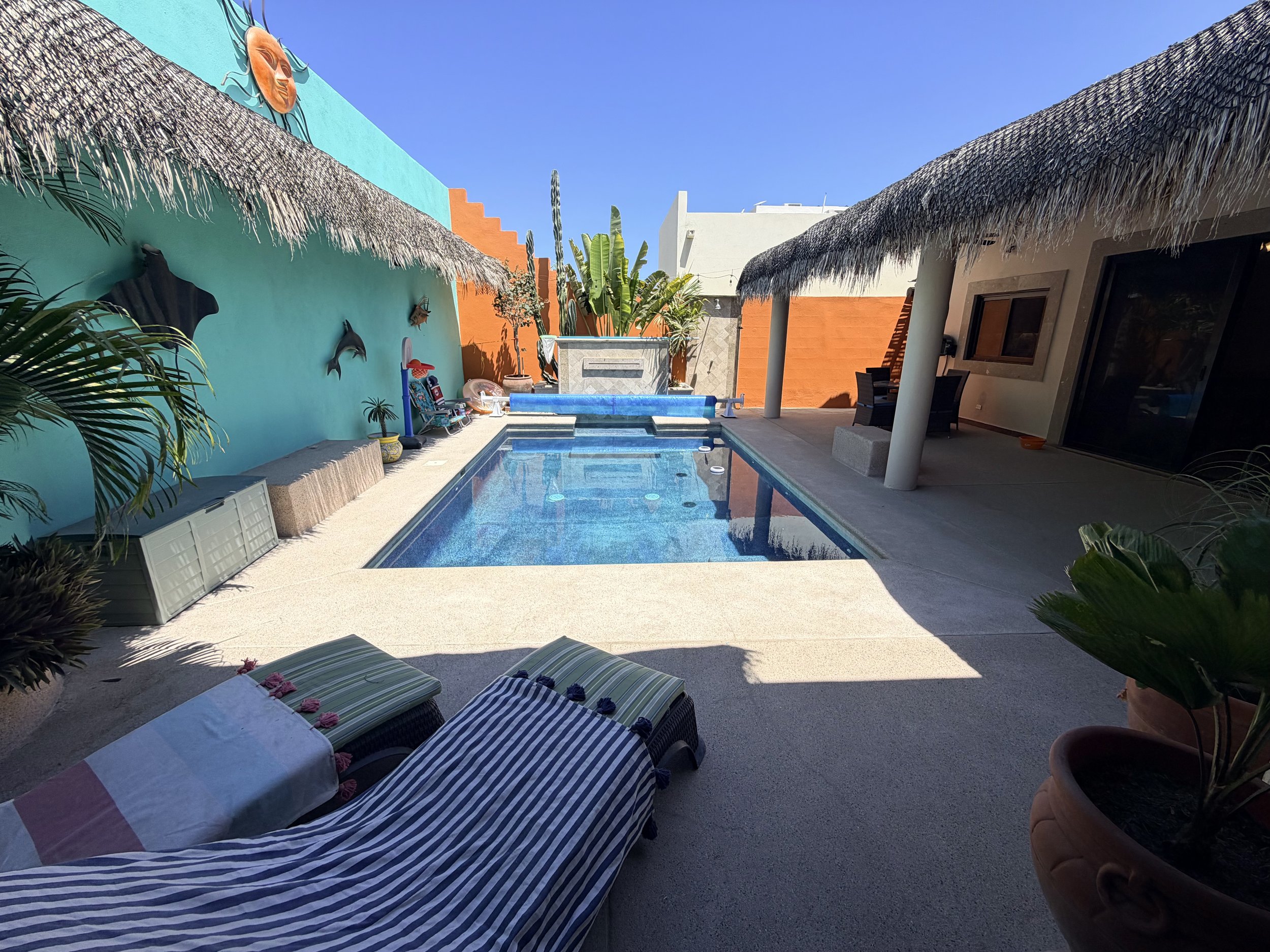 Poolside patio area with striped lounge chairs, a table with an umbrella, tropical plants, and a building with colorful walls and a thatched roof.