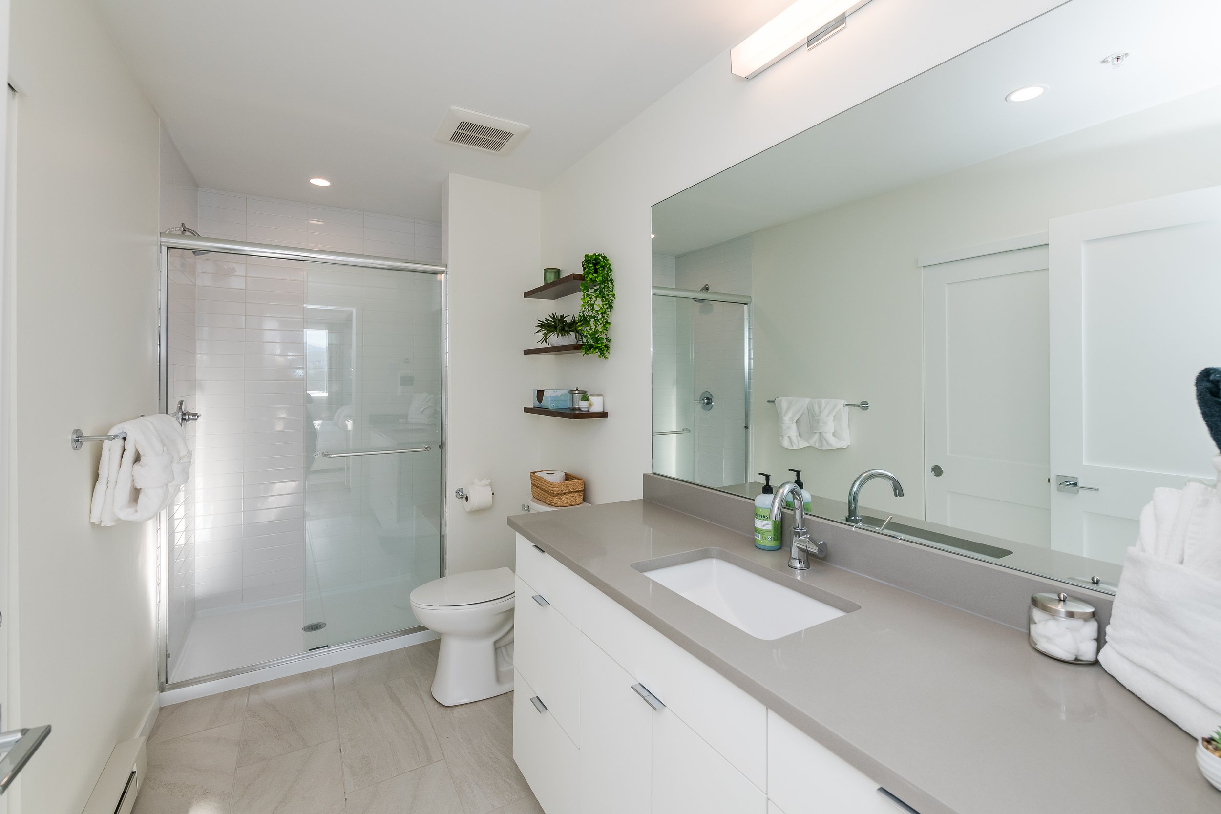 Modern bathroom with white walls, a large mirror, a sink with a counter, a toilet, and a shower with a glass door. Shelves with plants and toiletries, towels, and a basket are also visible.