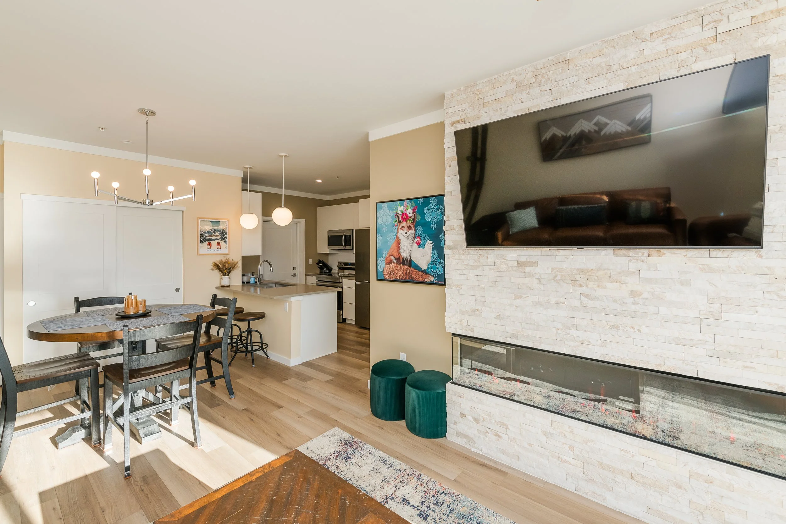 Open-concept living room and kitchen with a mounted flat-screen TV on a stone accent wall, a dining table with chairs, and artwork on the walls.