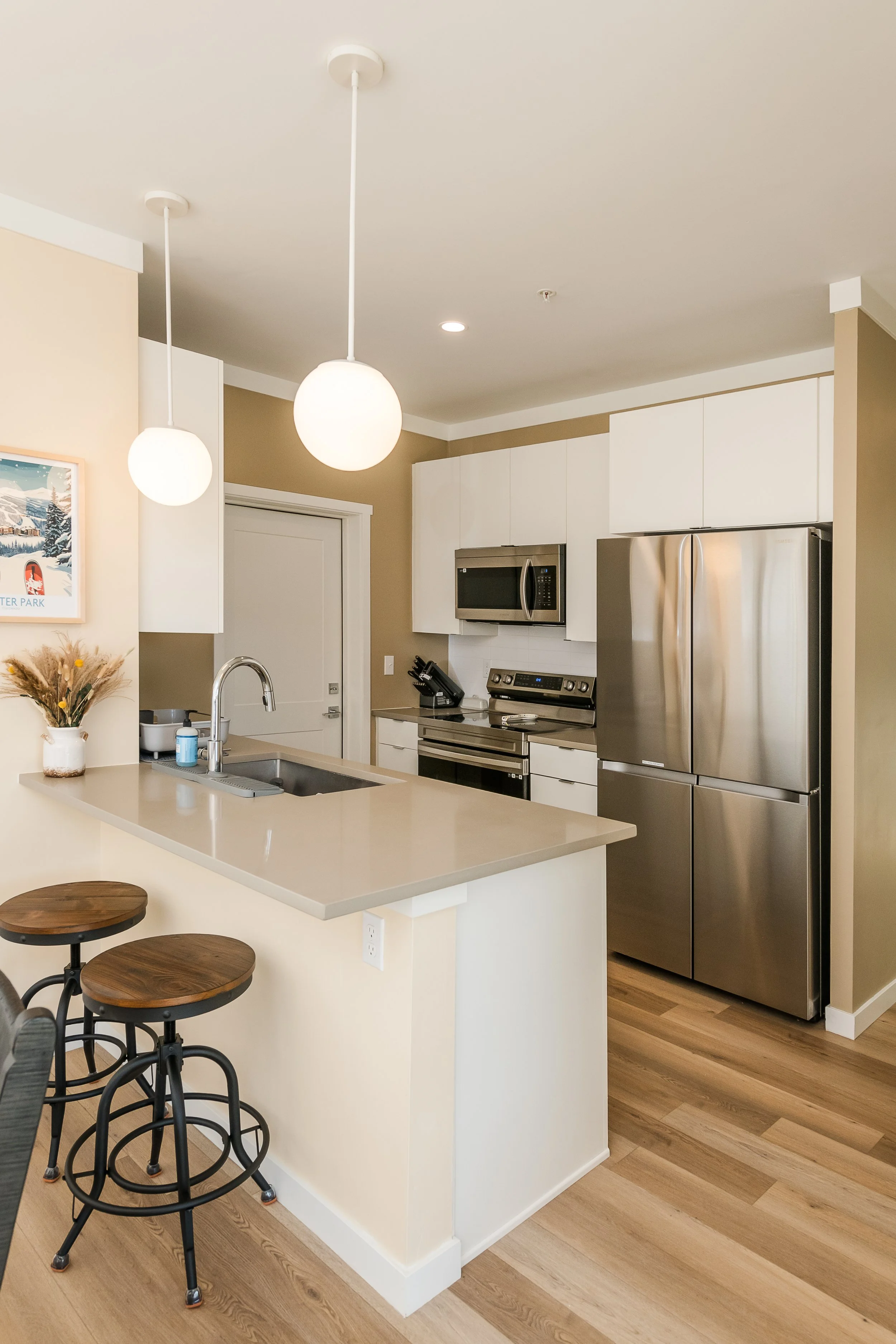 A modern kitchen with a beige and white color scheme. It features a white kitchen island with a light beige countertop, two wooden stools with black metal frames, and a sink with a silver faucet. The background shows white cabinets, a stainless steel