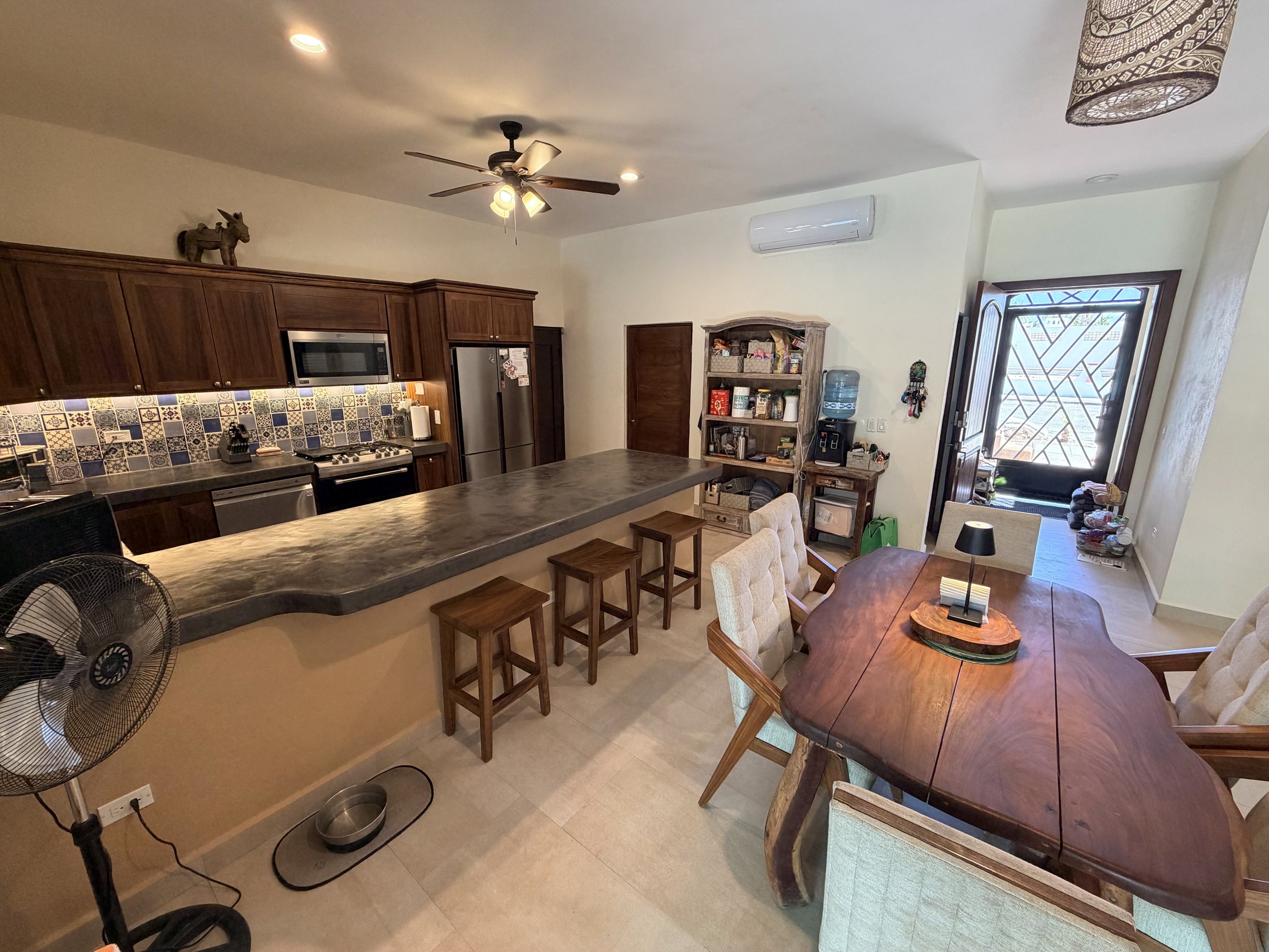 A kitchen and dining area with wooden cabinets, a ceiling fan, a water dispenser, and a doorway with a decorative door grille. There are chairs around a wooden dining table and a kitchen island with a dark countertop. The wall has a patterned tile ba