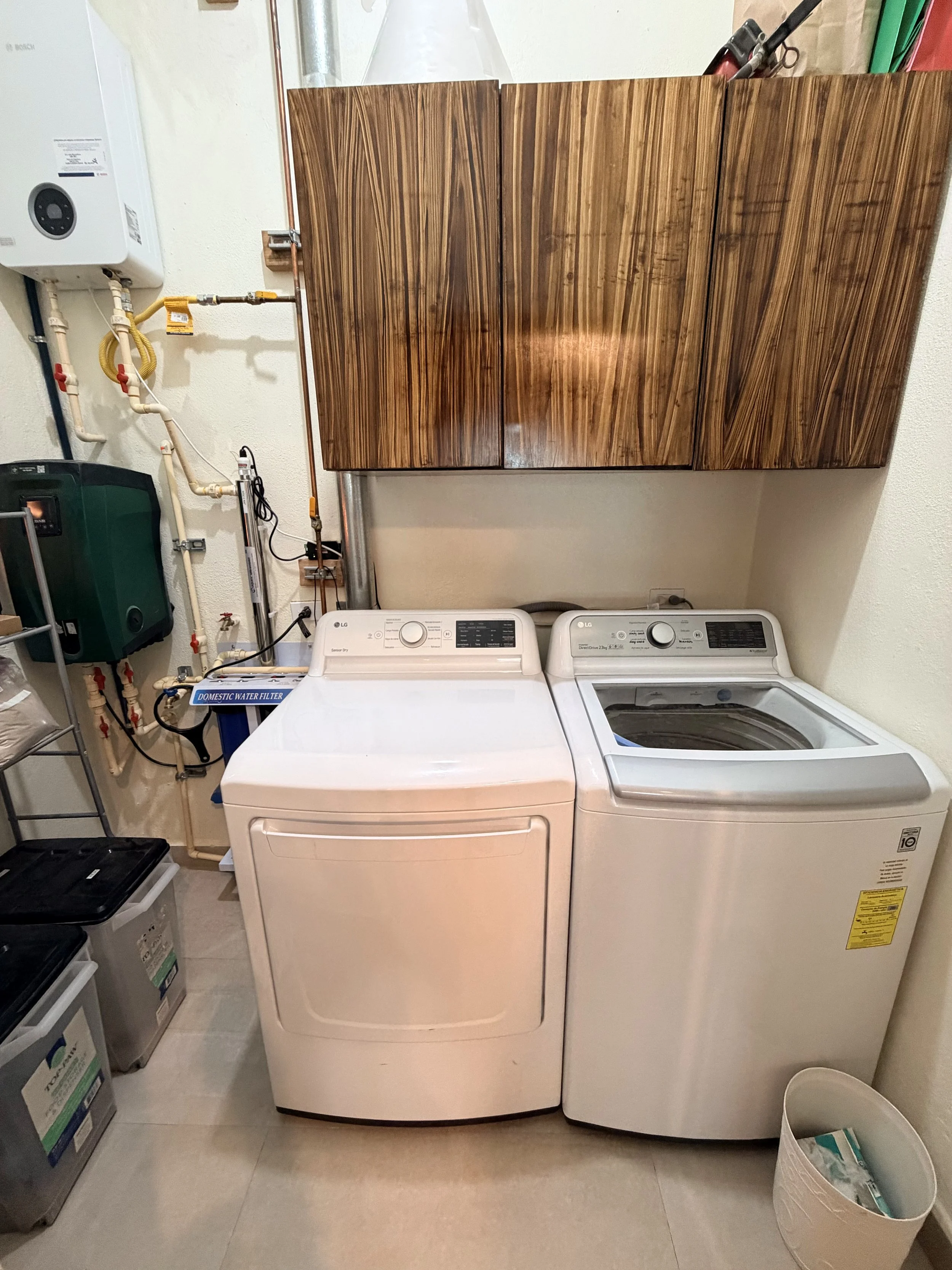 A laundry room featuring a white top-loading washer and dryer, wooden cabinets mounted on the wall, and items such as a trash bin and various storage boxes on the floor.