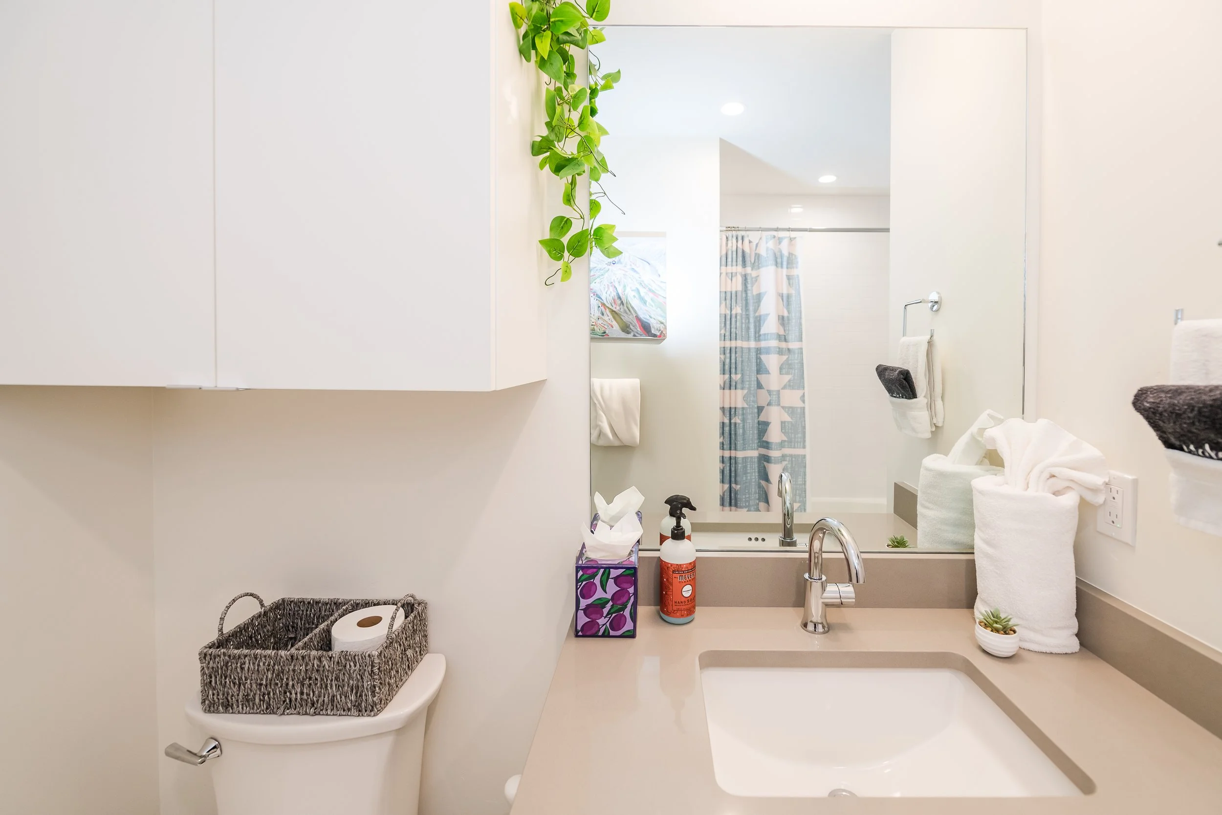 A clean and bright bathroom with a large mirror, a sink, and towels. There is a plant hanging on the wall above the mirror. The bathroom has a shower with a patterned curtain, a small colorful trash bin, and various toiletries and decorative plants o