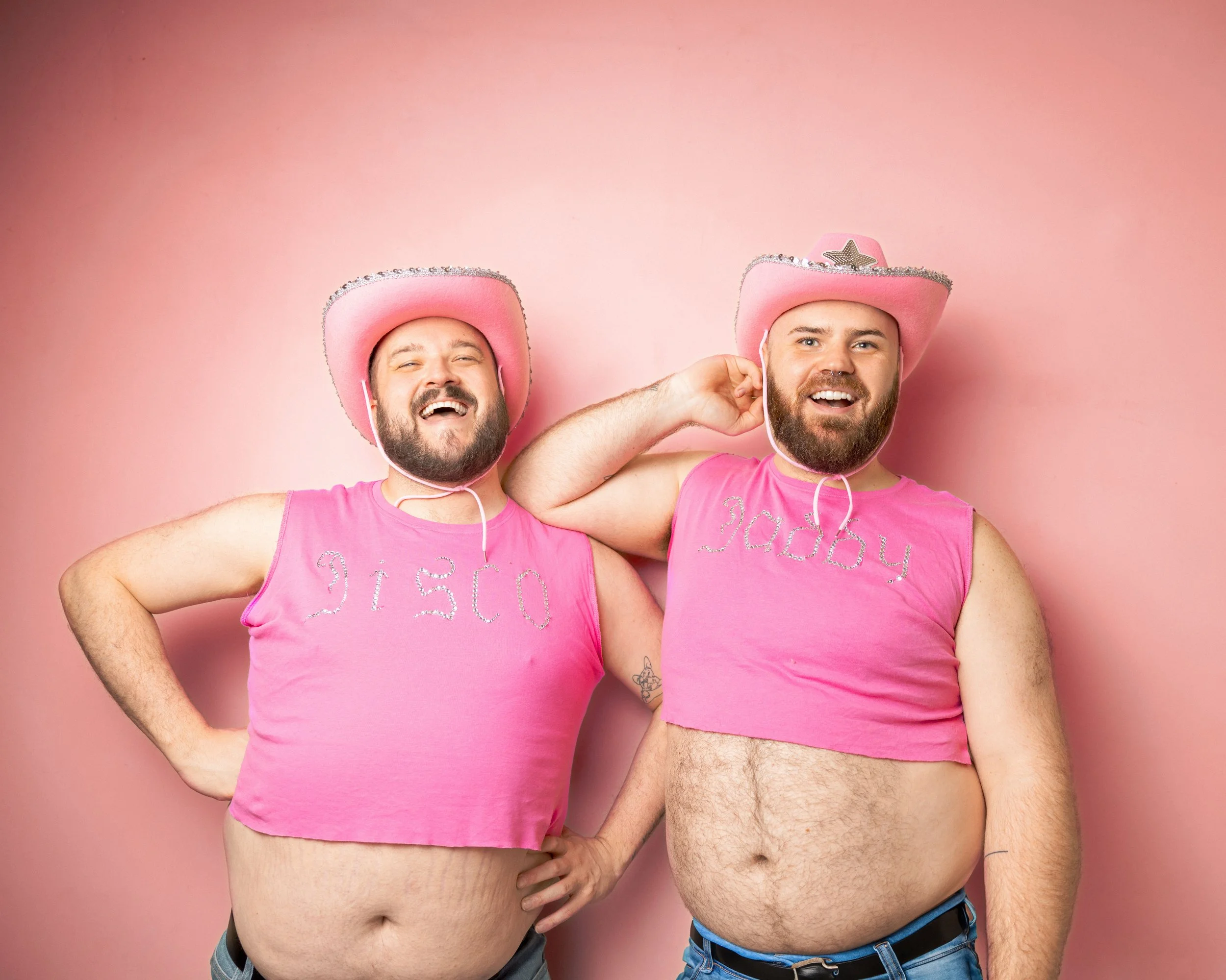 Two people with beards wearing pink sleeveless tops and pink cowboy hats, posing against a pink background. The man on the left has
