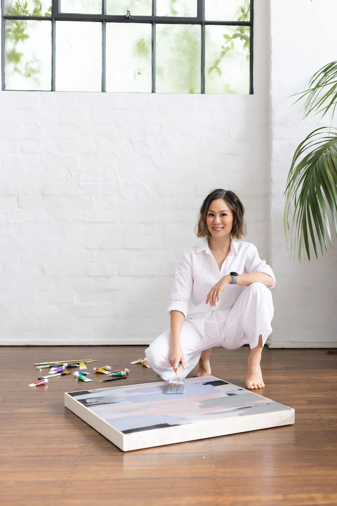 A woman in white clothes crouching on a wooden floor, holding a paintbrush, and painting a canvas with abstract art, with paints and brushes scattered around, in a bright room with white walls and a large window.