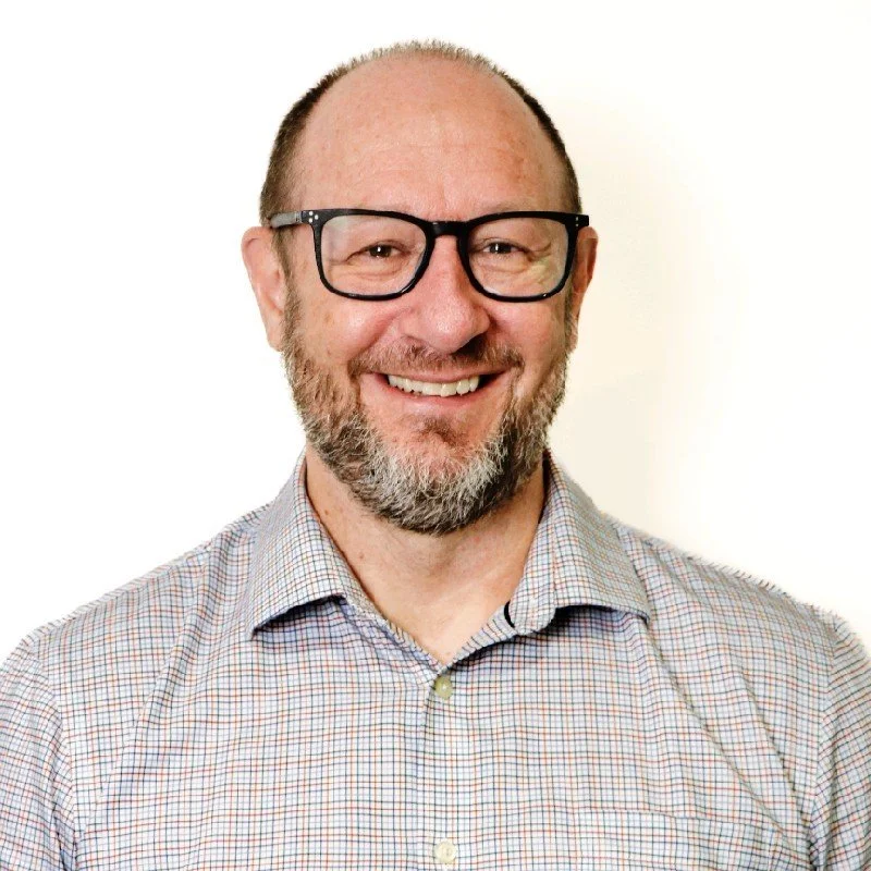 A smiling man with glasses and a beard, wearing a checked shirt against a white background.