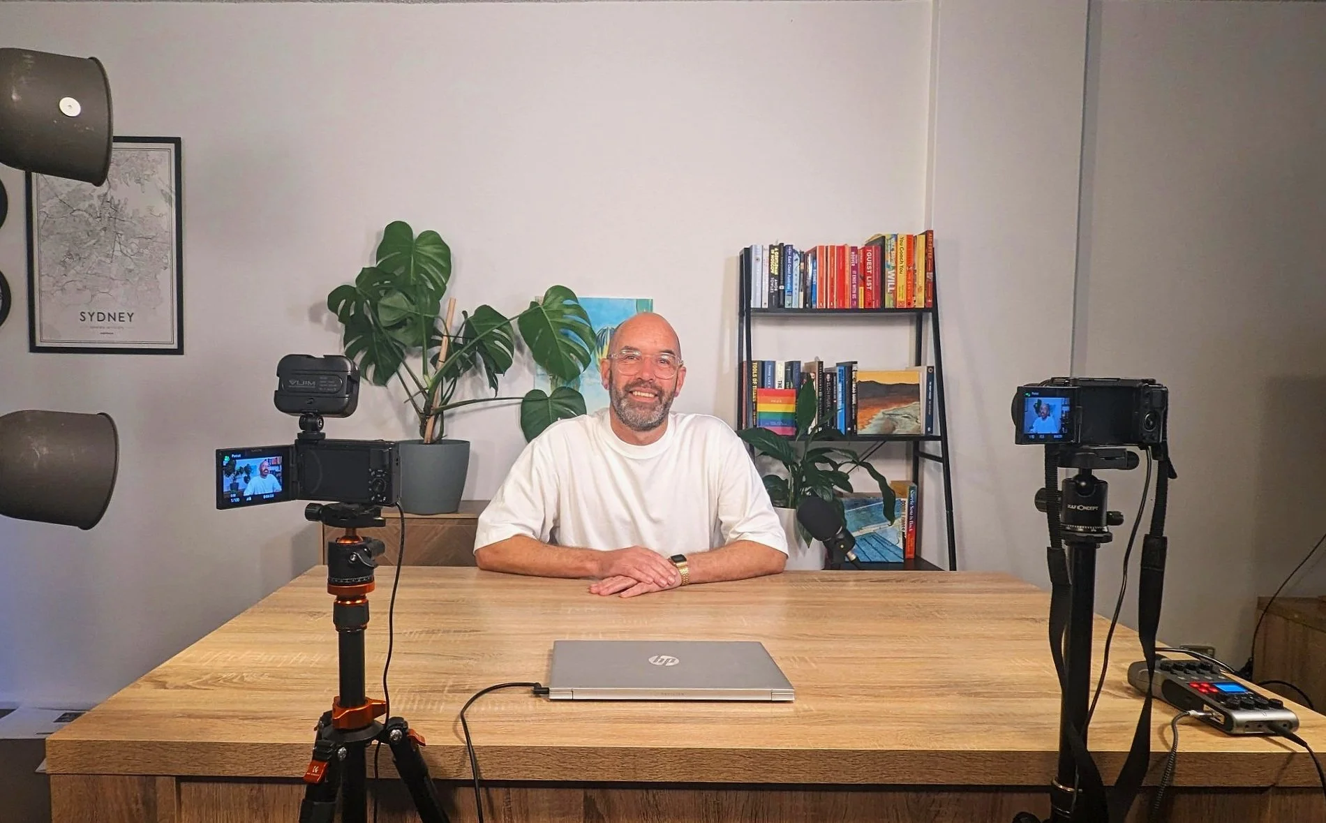 A man with glasses and a beard sitting at a wooden desk with a closed HP laptop, four cameras set up facing him, a microphone, a black device with red buttons, and a power strip. Behind him is a bookshelf with colorful books, large green plants, a framed map of Sydney, and a small painting of a landscape.