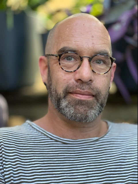 Man with shaved head and brown glasses, infront of a blurred background