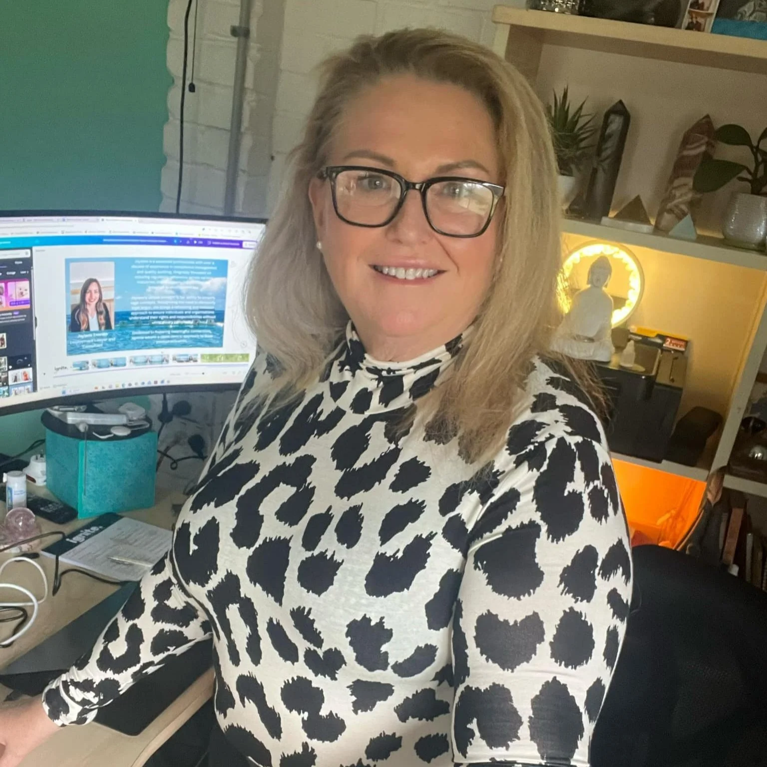 A woman with blonde hair and glasses smiling at the camera, sitting at a desk with a computer monitor displaying a webpage. She is wearing a black and white animal print top, and the background shows a cozy workspace with shelves, plants, and decorative items.