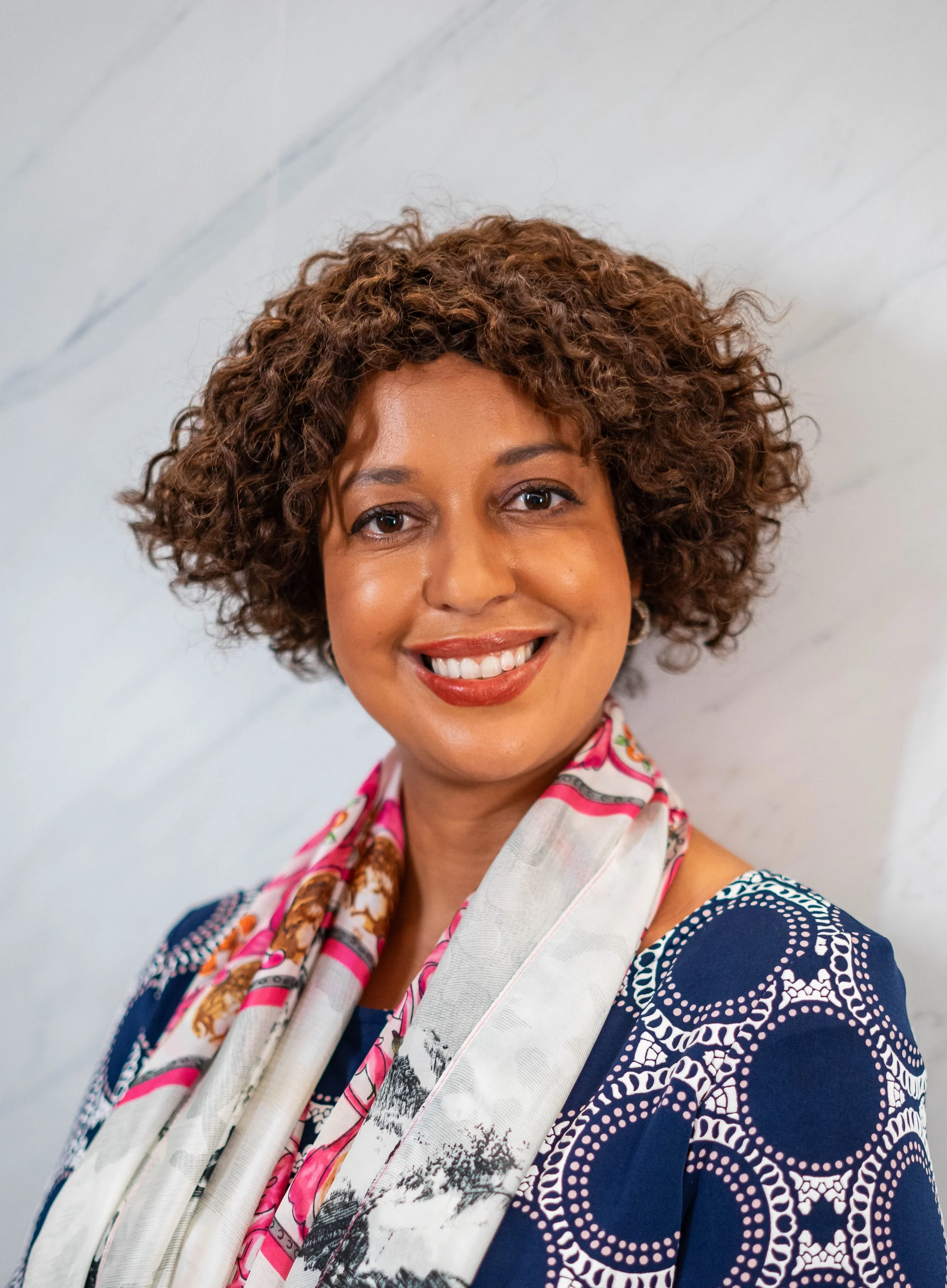A woman with short, curly brown hair smiling at the camera, wearing a patterned navy blue dress and a colorful scarf, standing against a light-colored wall.