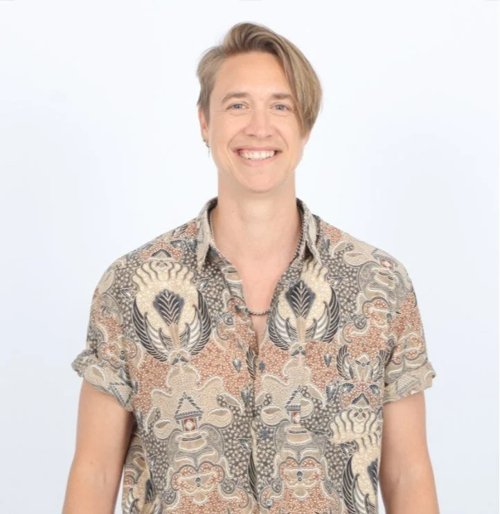 A smiling person with short blonde hair wearing a patterned short-sleeve shirt, standing against a plain white background.