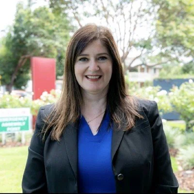 A woman with long brown hair smiling outdoors, wearing a black blazer over a blue blouse, with trees and a building in the background.