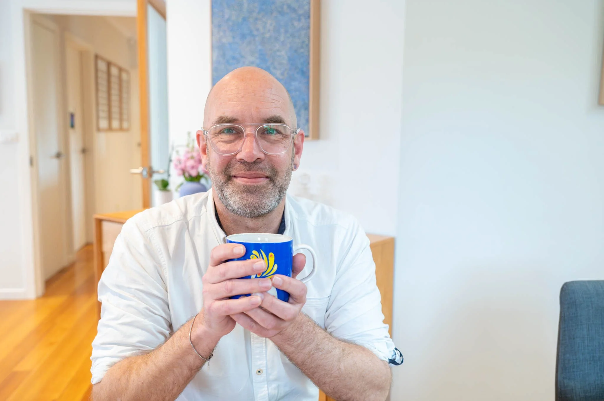 Man with a shaved head and glasses sat, with a mug in his hands, smiling