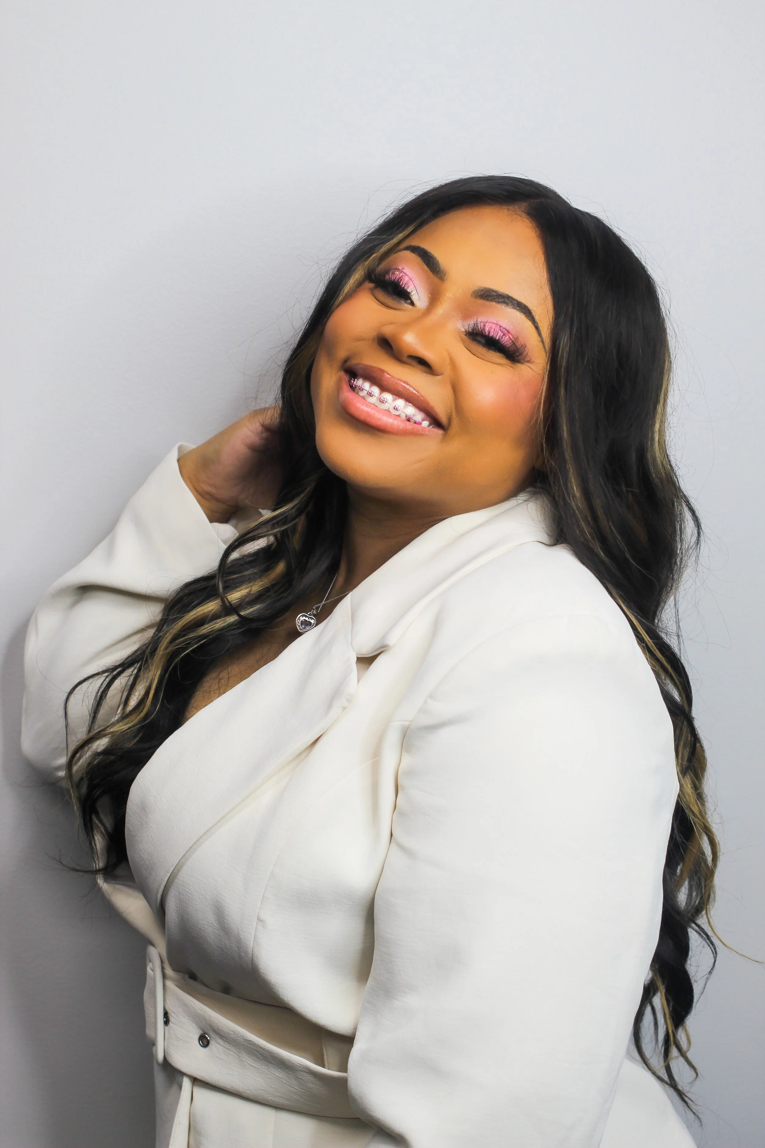 A woman smiling with closed eyes, wearing pink eyeshadow, a white blazer, and braces, standing against a plain light gray wall.