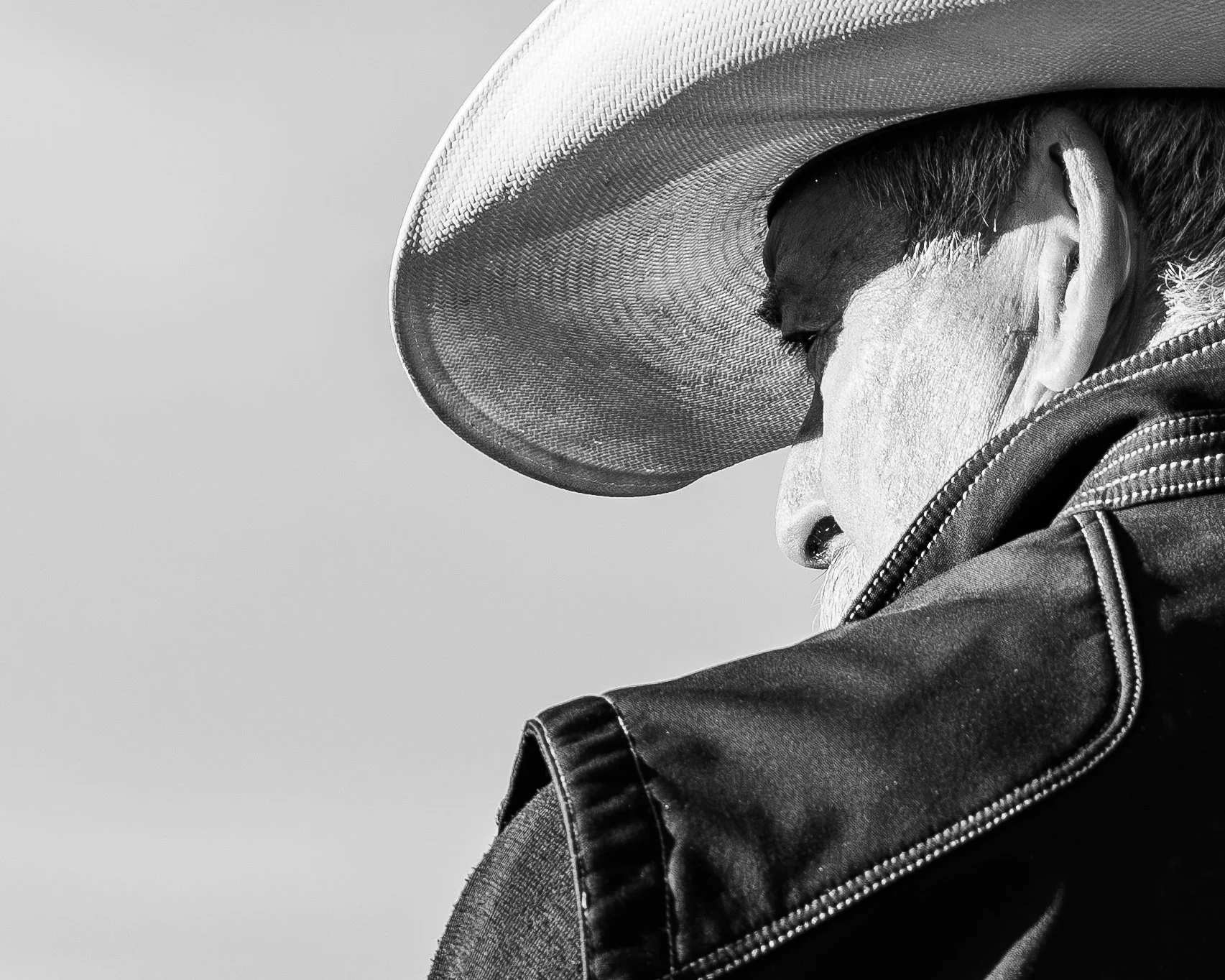 Side view of a man wearing a large cowboy hat, sunglasses, a jacket, and a shirt, looking downward with a serious expression.