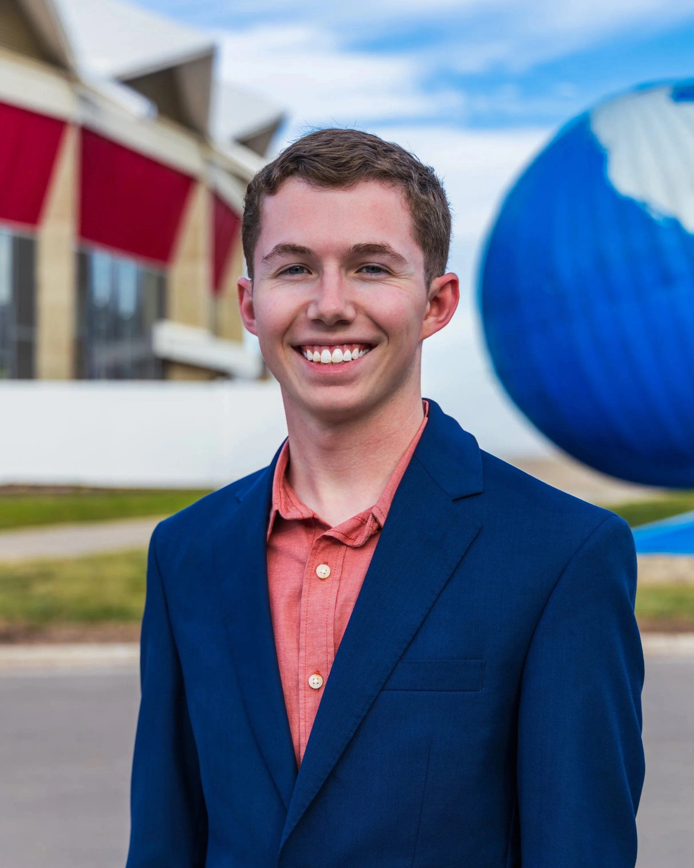 A young man(Cade Claycomb) in a blue blazer and salmon-colored shirt smiling outdoors with a blue sky and a modern building in the background.