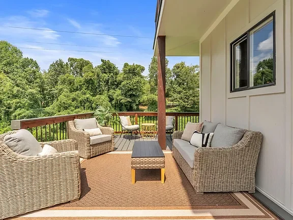 Outdoor patio with wicker furniture, including two armchairs, a loveseat, and a coffee table, on a brown rug. The patio has a wooden railing and overlooks a lush green landscape with trees under a blue sky.