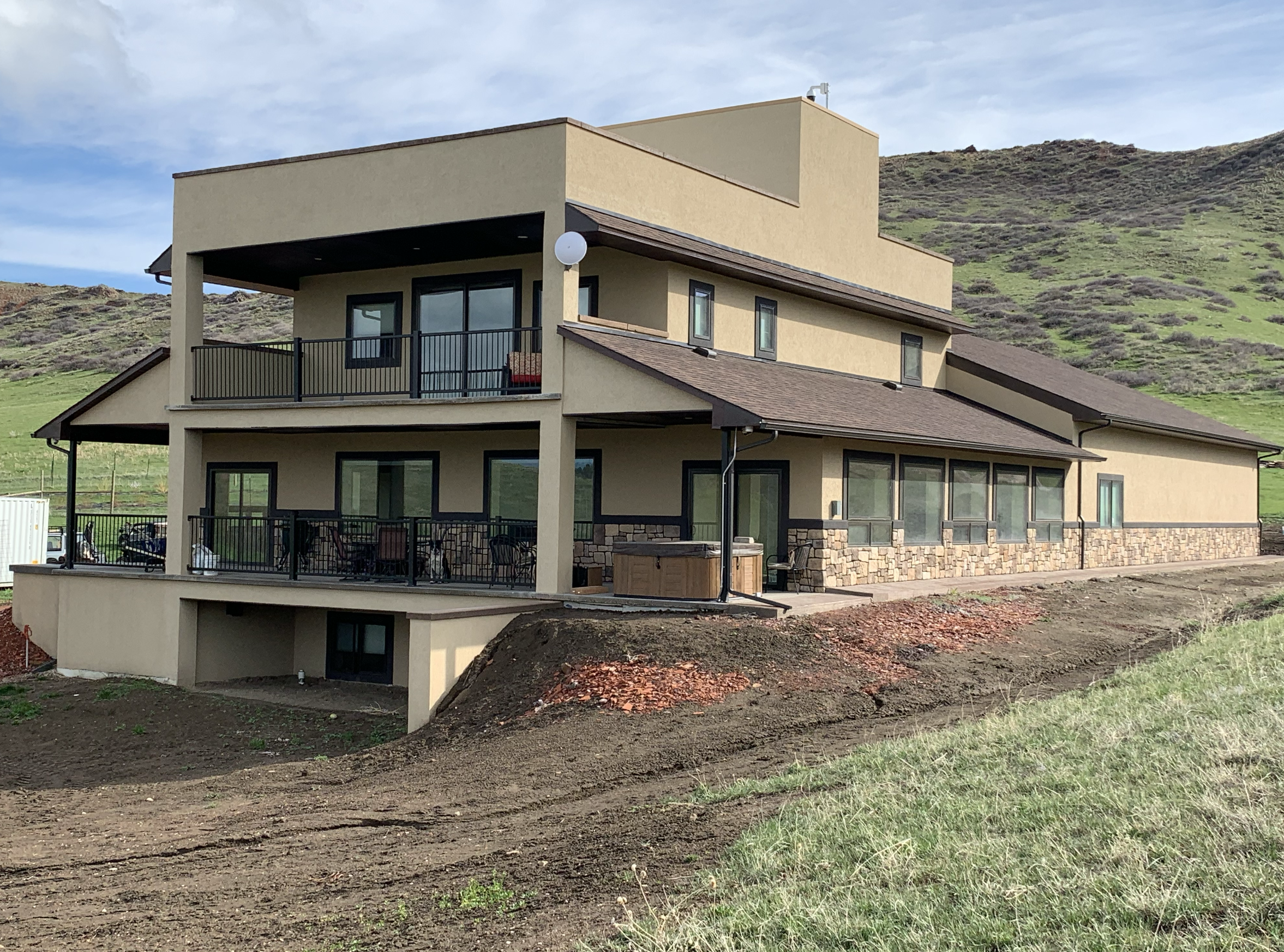 Modern house with multiple levels, beige exterior walls, brown roofs, large windows, and a black balcony railing, situated on a hillside with grassy and rocky terrain beneath.