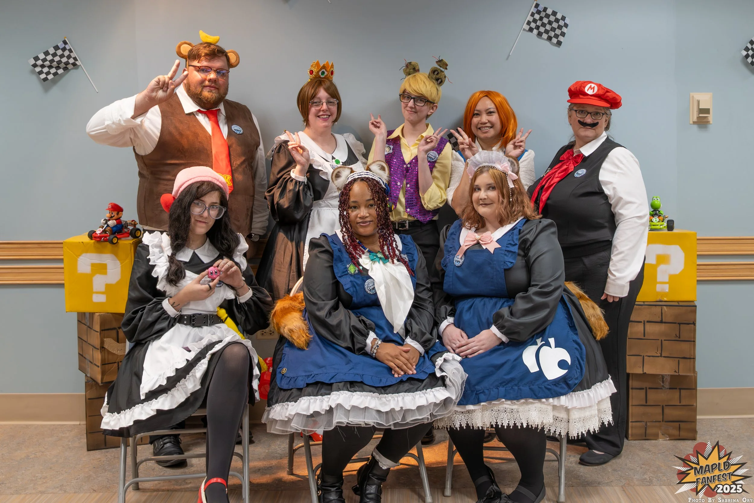 Group of nine people dressed in costumes, some with animal accessories, posing with peace and victory signs in front of a blue wall with checkered flags and Mario-themed toys, at an event called MAPLD Fan Fest 2025.