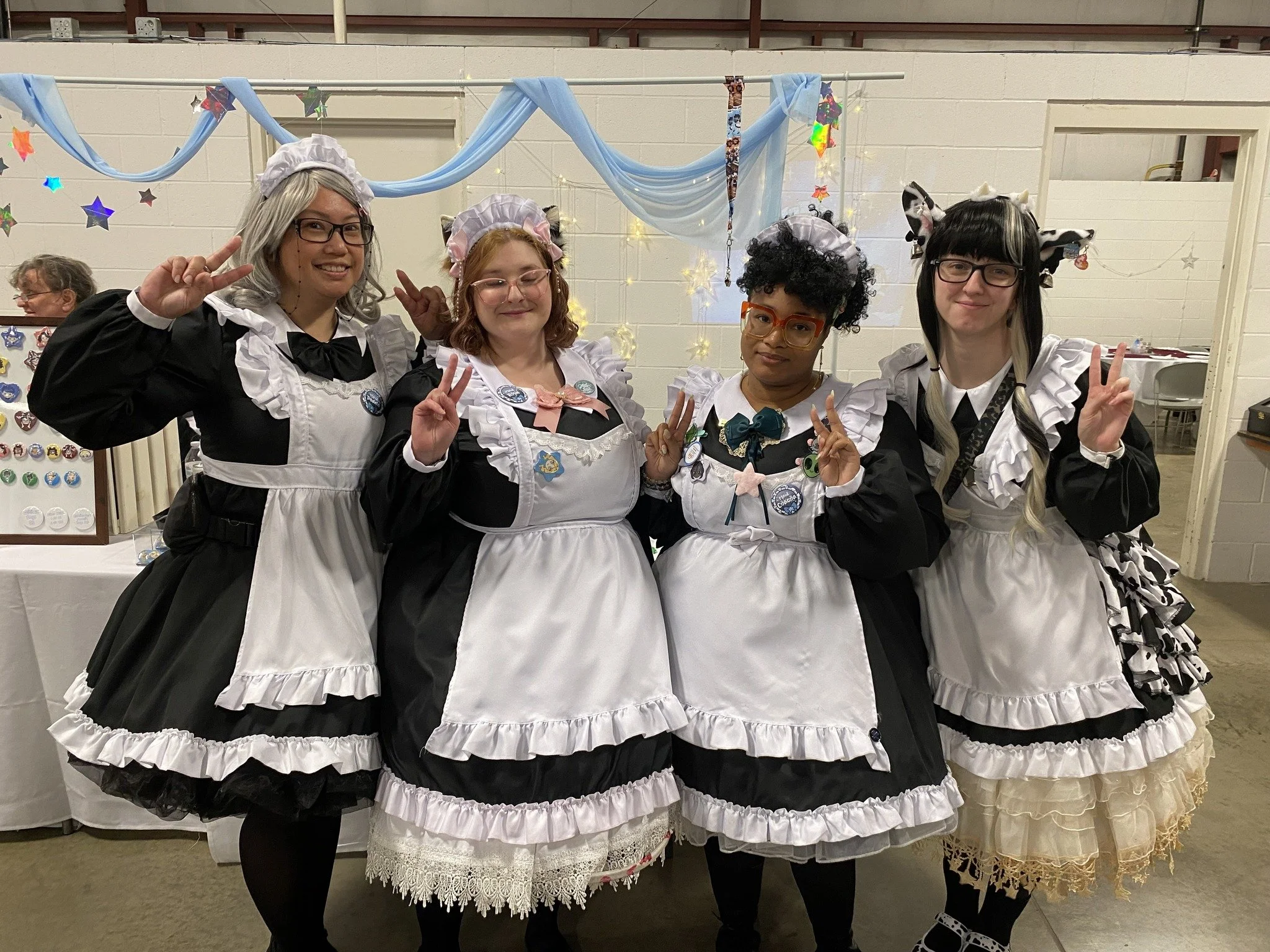 Four women dressed in maid costumes with frilly black and white dresses, headpieces, and accessories, posing and smiling at a party with decorative star and star-shaped balloon backdrop.