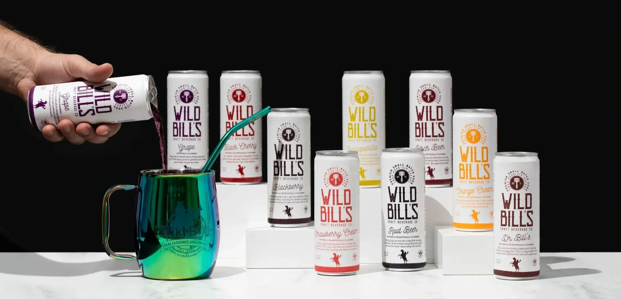 A hand pouring purple liquid into a rainbow-colored mug with a straw, with multiple cans of Wild Bill's craft beverages arranged behind on a white surface against a black background.