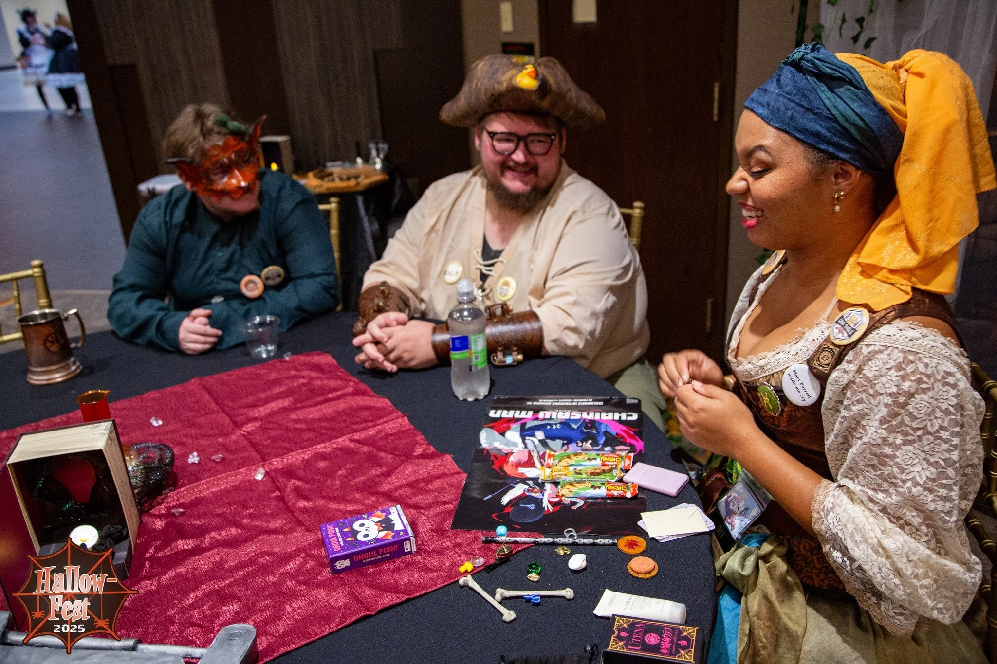 Three people in costumes sit at a table during Hallow Fest 2025. The person in the middle wears glasses and a pirate hat with a yellow rubber duck on top. The person on the left has red face paint and green hair, while the person on the right wears a colorful head wrap and a lace dress. The table is decorated with Halloween themed items, including candy, dice, and decorations.