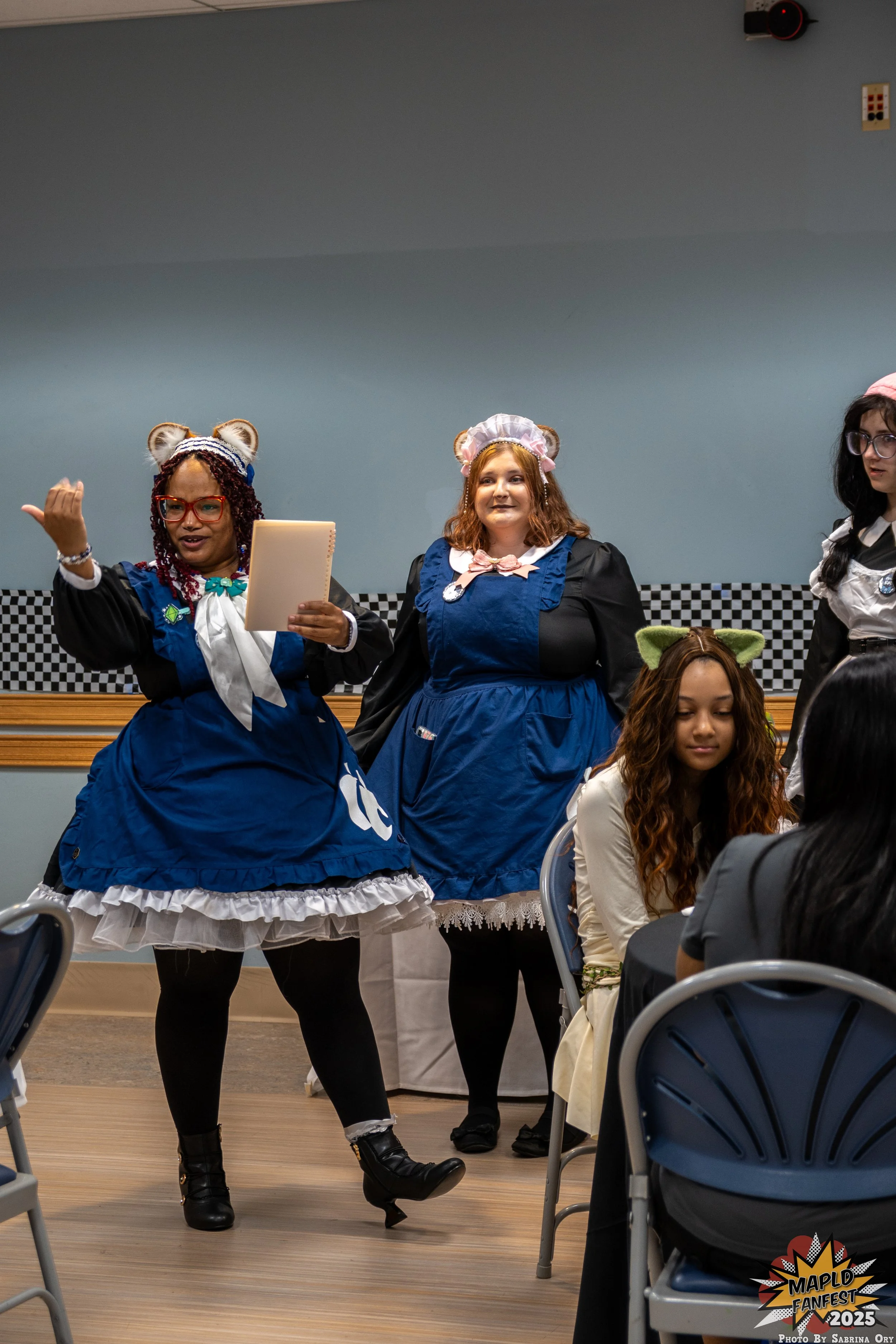 Three women dressed in cosplay costumes standing behind a table at an indoor event, with some sitting in chairs in front of them. One woman is wearing a blue dress with a white apron, a pink bonnet, and has red hair. The second woman is wearing a similar blue dress with a pink bonnet and has brown hair. The third woman is wearing a maid costume with glasses and has long, dark hair. The setting appears to be a convention or fan event, with a checkered wall decoration and a logo in the bottom right corner that says "Maple Fanfest 2025".