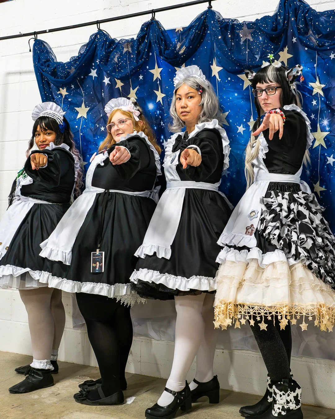 Four women dressed as maids in black and white uniforms standing in a row, pointing forward, with a starry blue backdrop behind them.