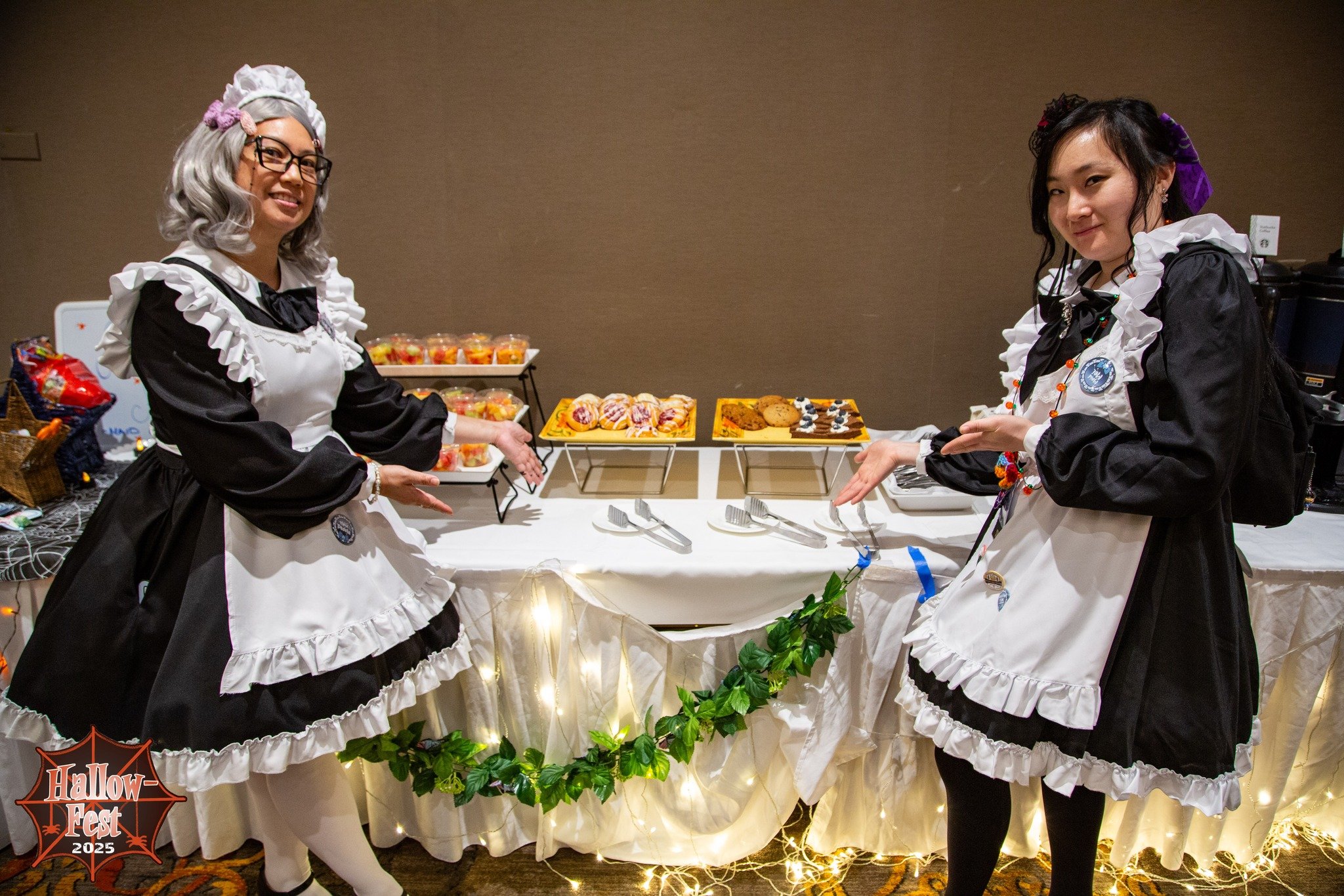 Two women dressed in black and white maid costumes with frilled aprons and headbands stand at a dessert buffet table. The table is decorated with string lights and green foliage, and has an assortment of desserts, including cookies and fruit cups. One woman has gray hair with pink hair rollers and glasses, and the other has black hair tied with purple and red accessories. The background is a plain beige wall.