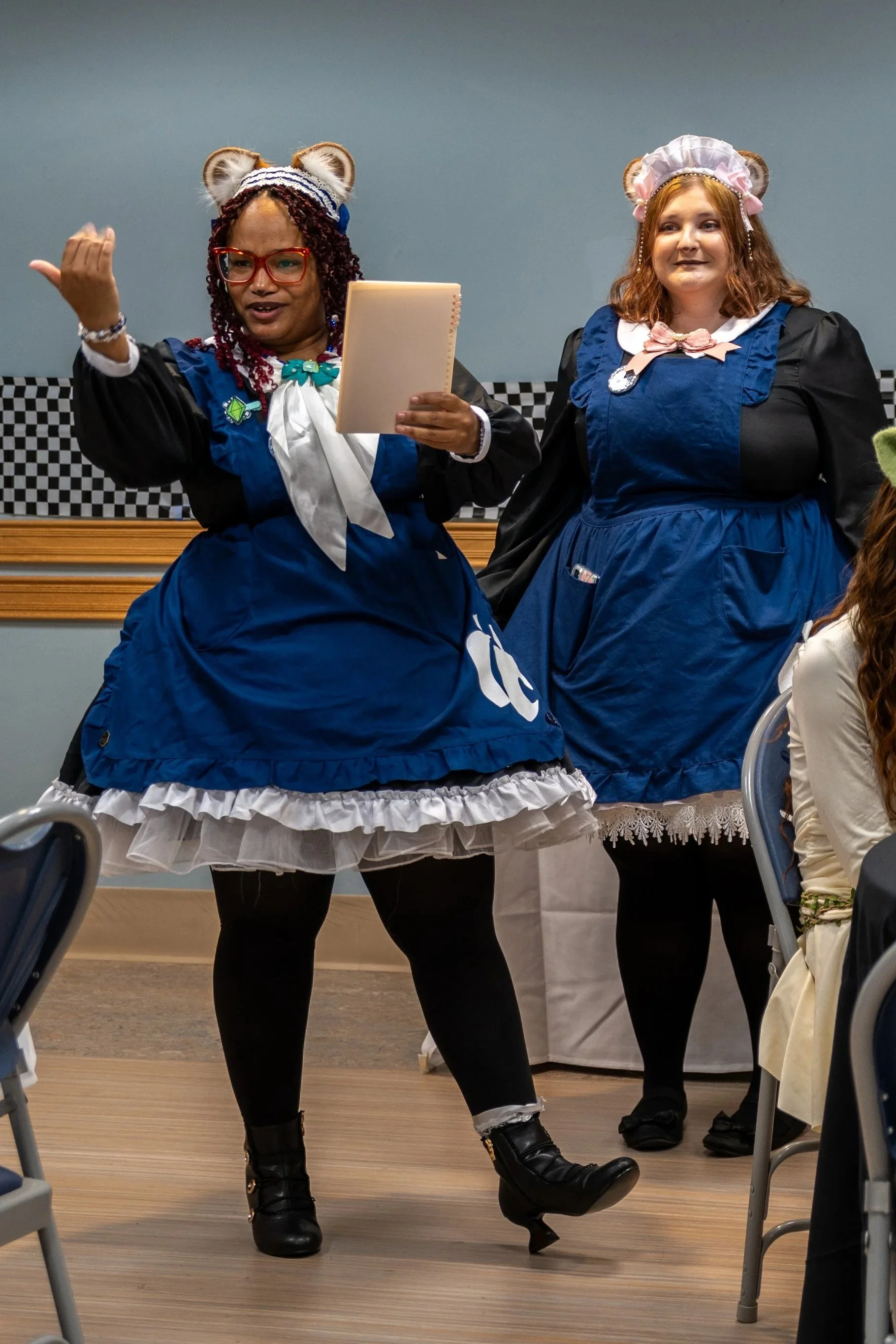 Two women dressed in Victorian maid costumes stand indoors, engaging with an audience. The woman on the left wears a blue dress with a white apron, red glasses, and a headband with animal ears, holding a notebook. The woman on the right wears a similar blue dress with a pink bow and a headpiece with animal ears. The background features a gray wall with checkered trim.