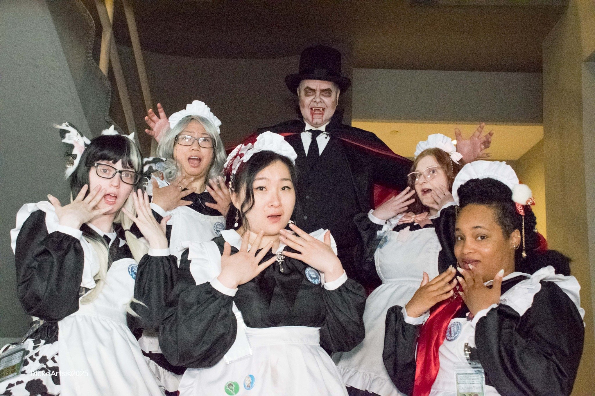 Group of people dressed in Halloween costumes posing together, with one dressed as a vampire and others as maids or nurses, indoors.