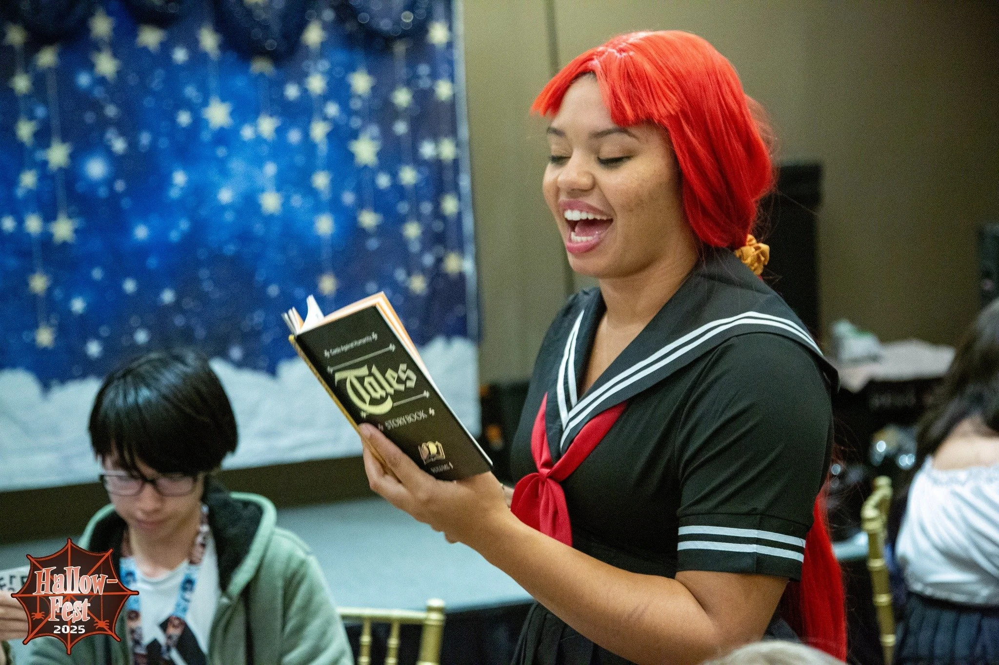 A woman with bright red hair styled with bangs, wearing a black and white sailor-style outfit with a red necktie, is smiling and reading a booklet titled 'Tales' at a Halloween event in 2025, with a blue starry Halloween decoration in the background.