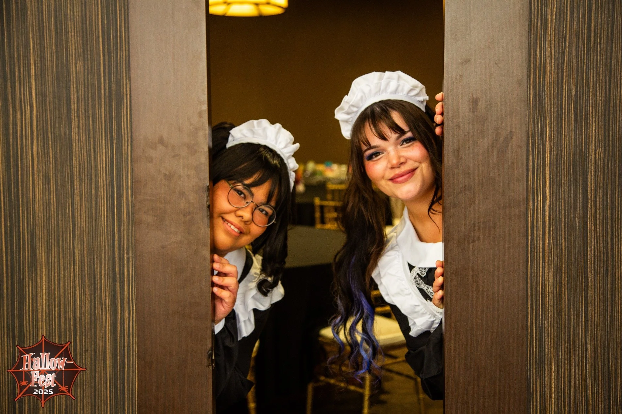 Two women dressed as waitresses peek through a small opening in a door at a Halloween event in 2025.