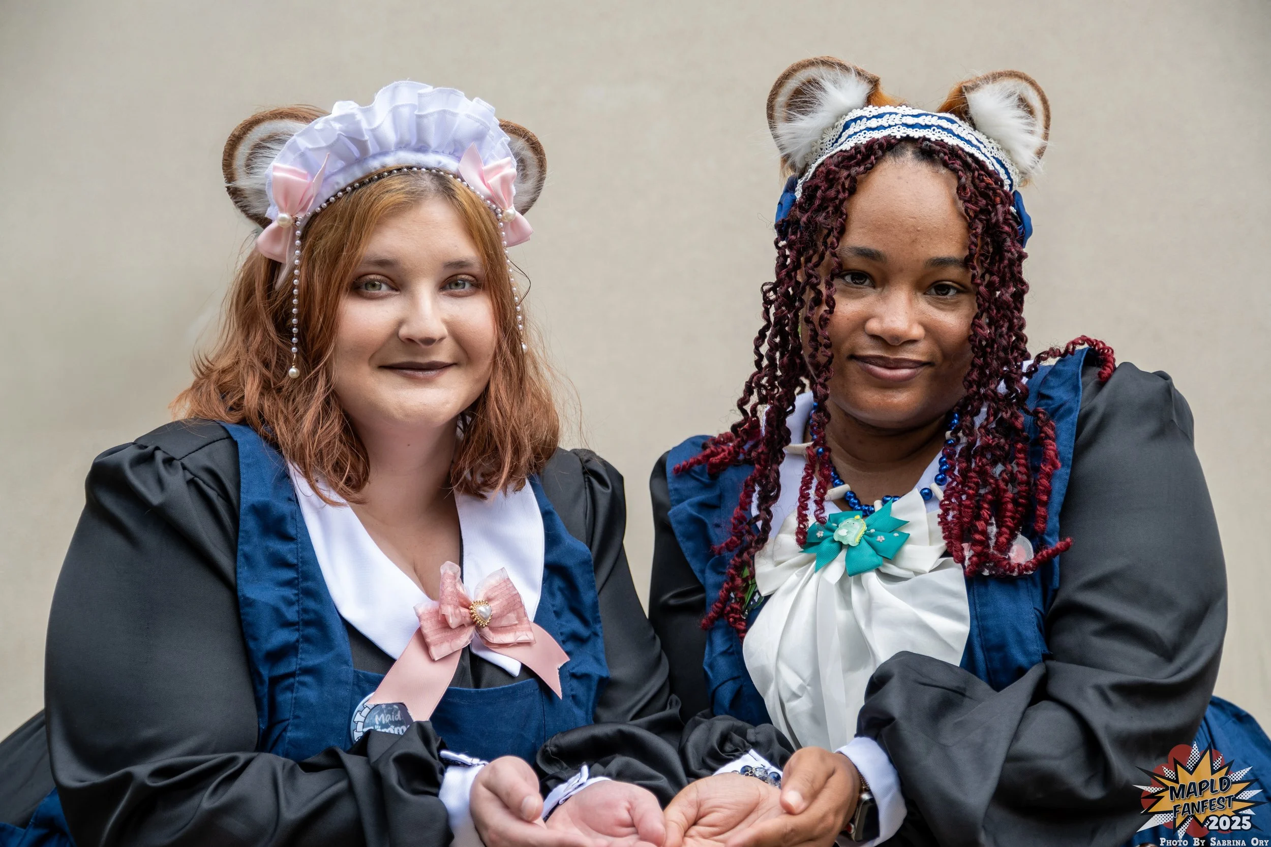 Two women dressed in cosplay costumes with animal ear headbands and colorful necklaces, smiling at the camera at an event. One has red hair and the other has dark, curly hair.