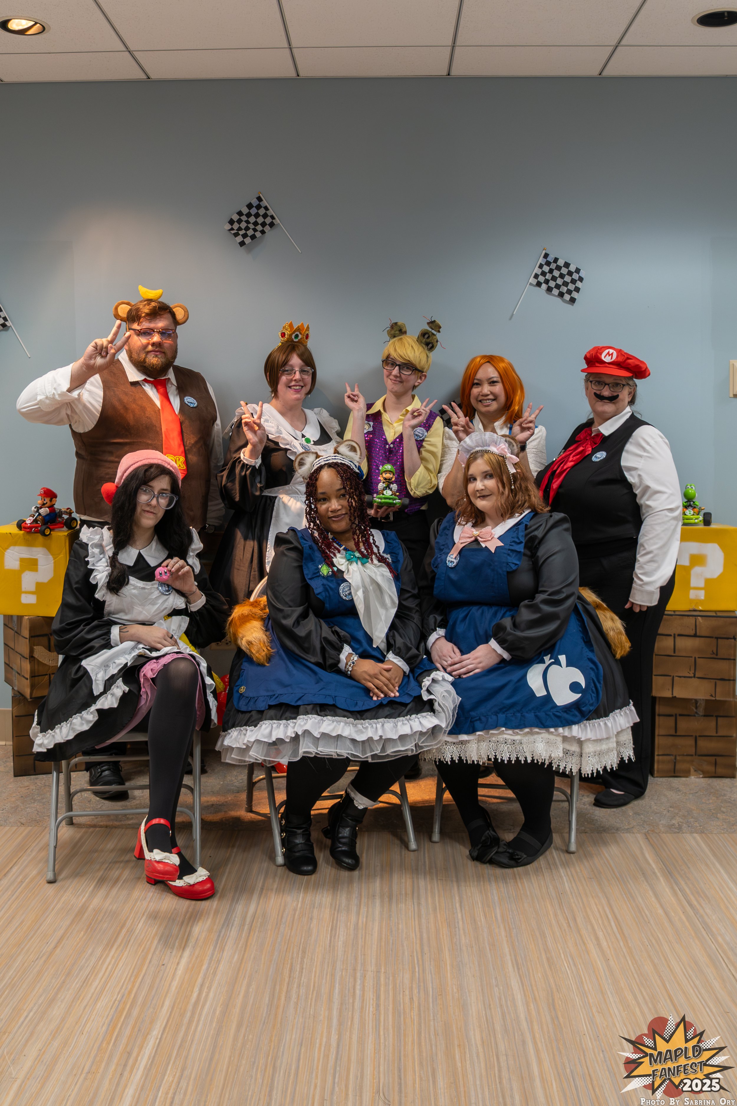 A group of people dressed in costumes posing at an indoor event, with some holding peace signs and others with props. Background includes checkered flags and a table with Mario-themed decorations, and a photo booth backdrop featuring a logo that says "Maple Fanfest 2025".