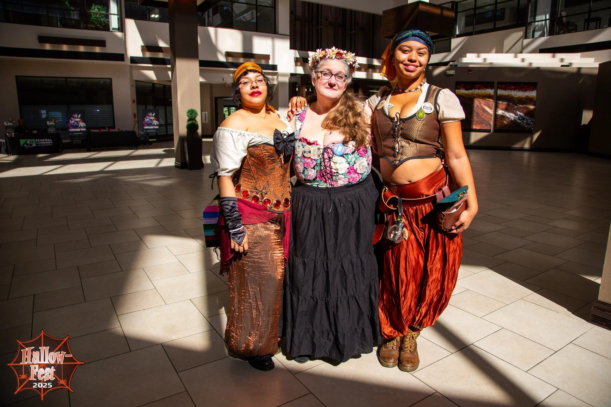 Three women dressed in Halloween costumes standing together inside a modern building with sunlight streaming through large windows. The woman on the left wears a vintage-style dress with a corset, gloves, and a headscarf. The woman in the middle wears a floral blouse and a flower crown. The woman on the right is dressed in a medieval or fantasy-inspired outfit with a headscarf, corset, and leather accessories. There is a logo in the bottom left corner that reads 'Hallow-Fest 2025'.