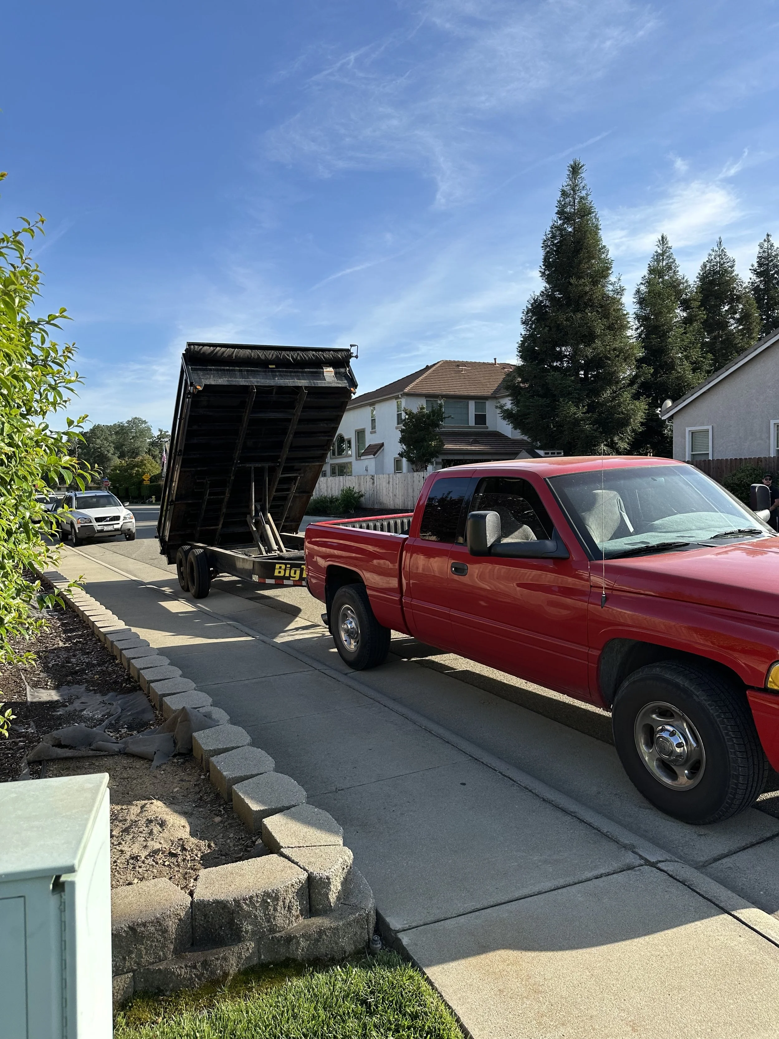 A red pickup truck with a raised flatbed parked on a driveway, with other vehicles and a residential house with a brown roof and trees in the background, under a clear blue sky.