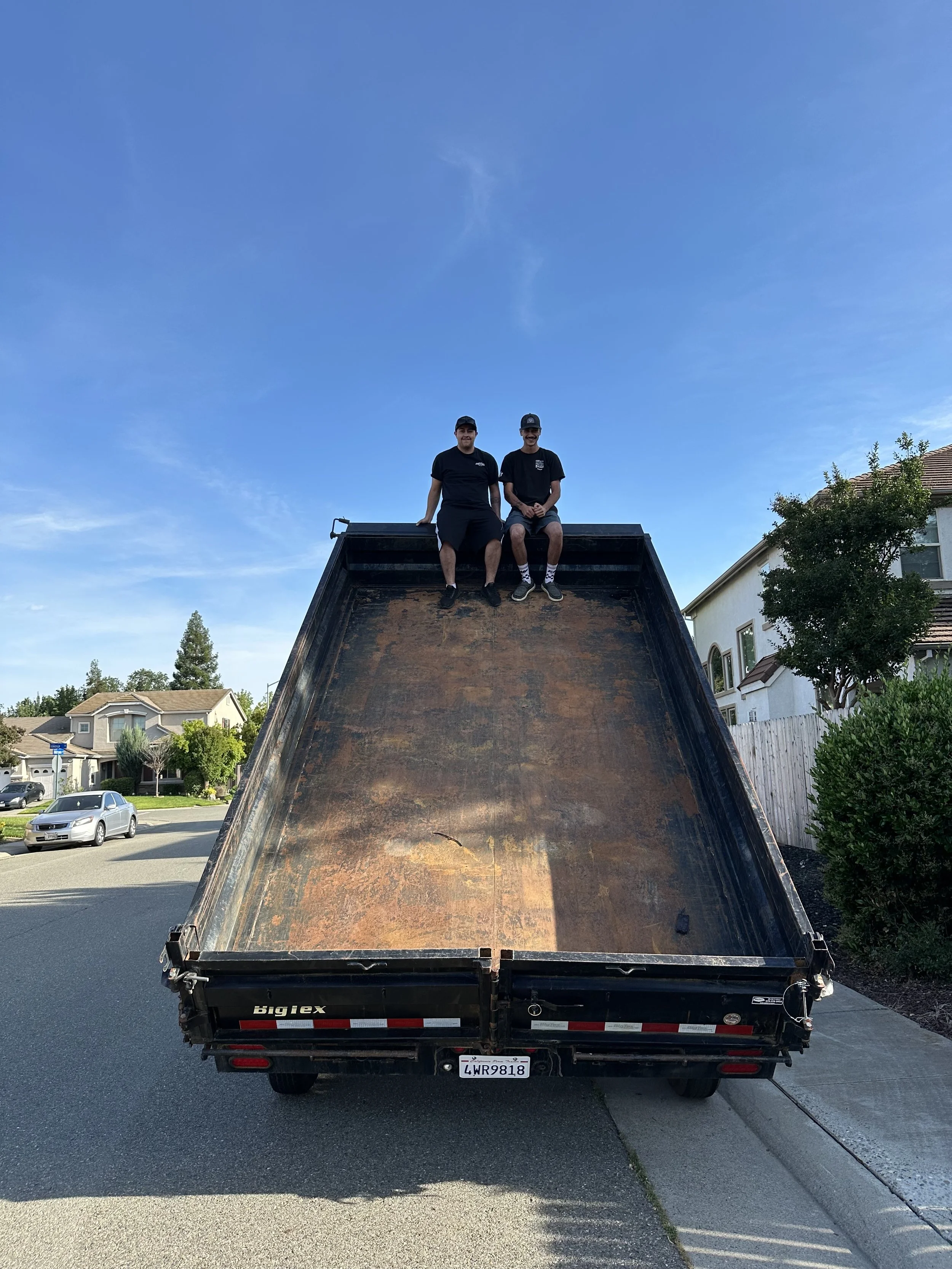 Two men sit on the edge of a large black truck bed in a suburban neighborhood under a clear blue sky. They are smiling and wearing casual clothing and caps, with trees and houses visible in the background.