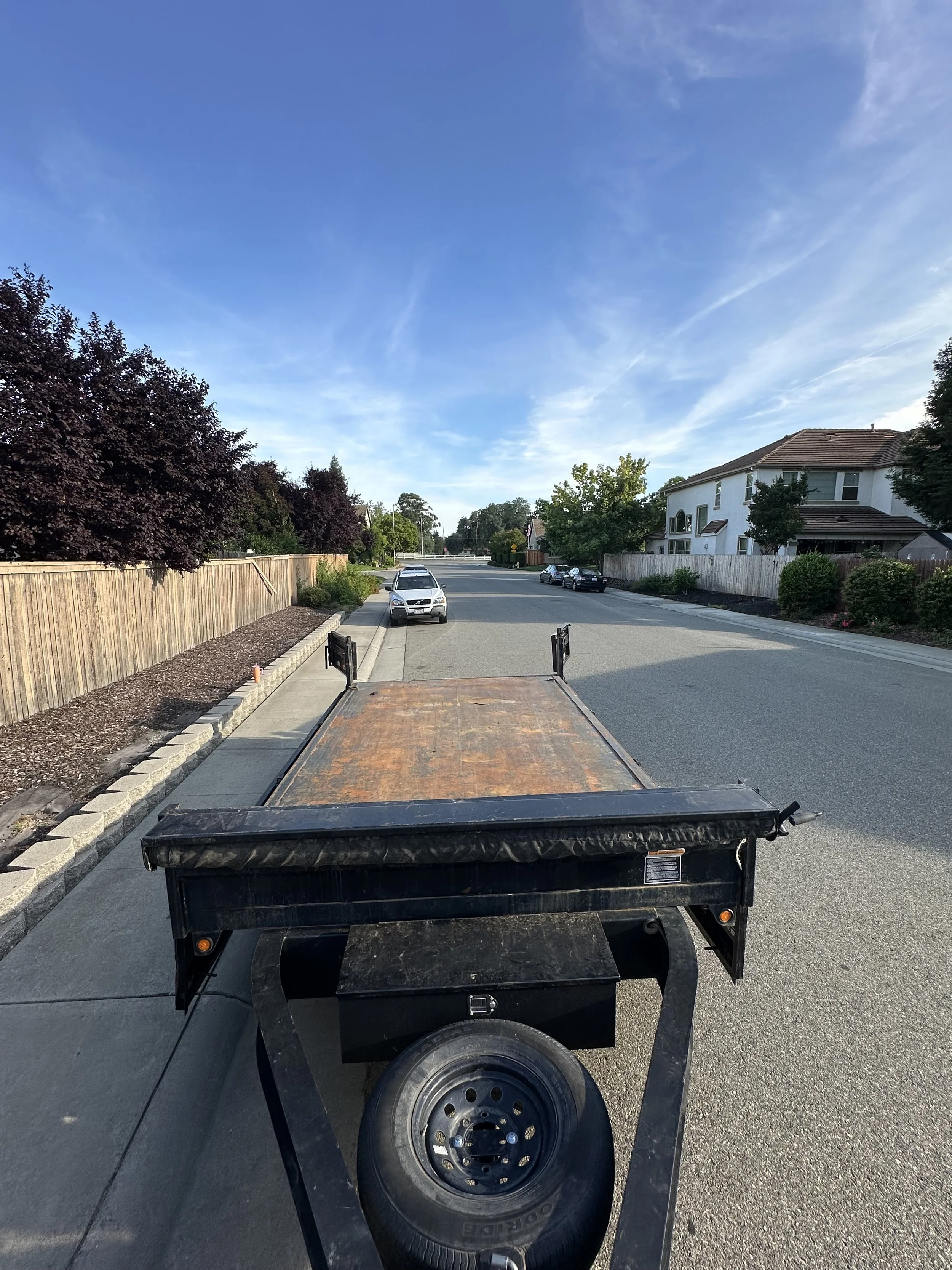 A black utility trailer parked on a residential street, with a rusty wooden flatbed and black metal sides. Several houses and trees line the street, and there is a blue sky with some clouds overhead.