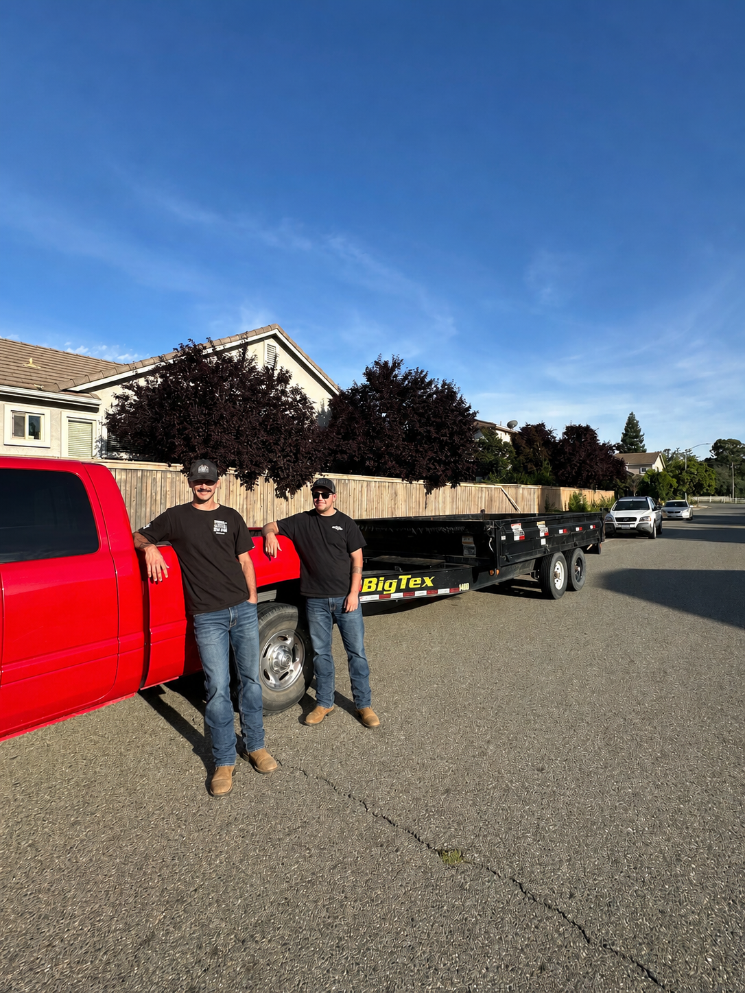 Two men in black t-shirts and jeans standing near a red pickup truck with a black flatbed trailer attached, under a tree with purple foliage, in a suburban neighborhood on a clear day.