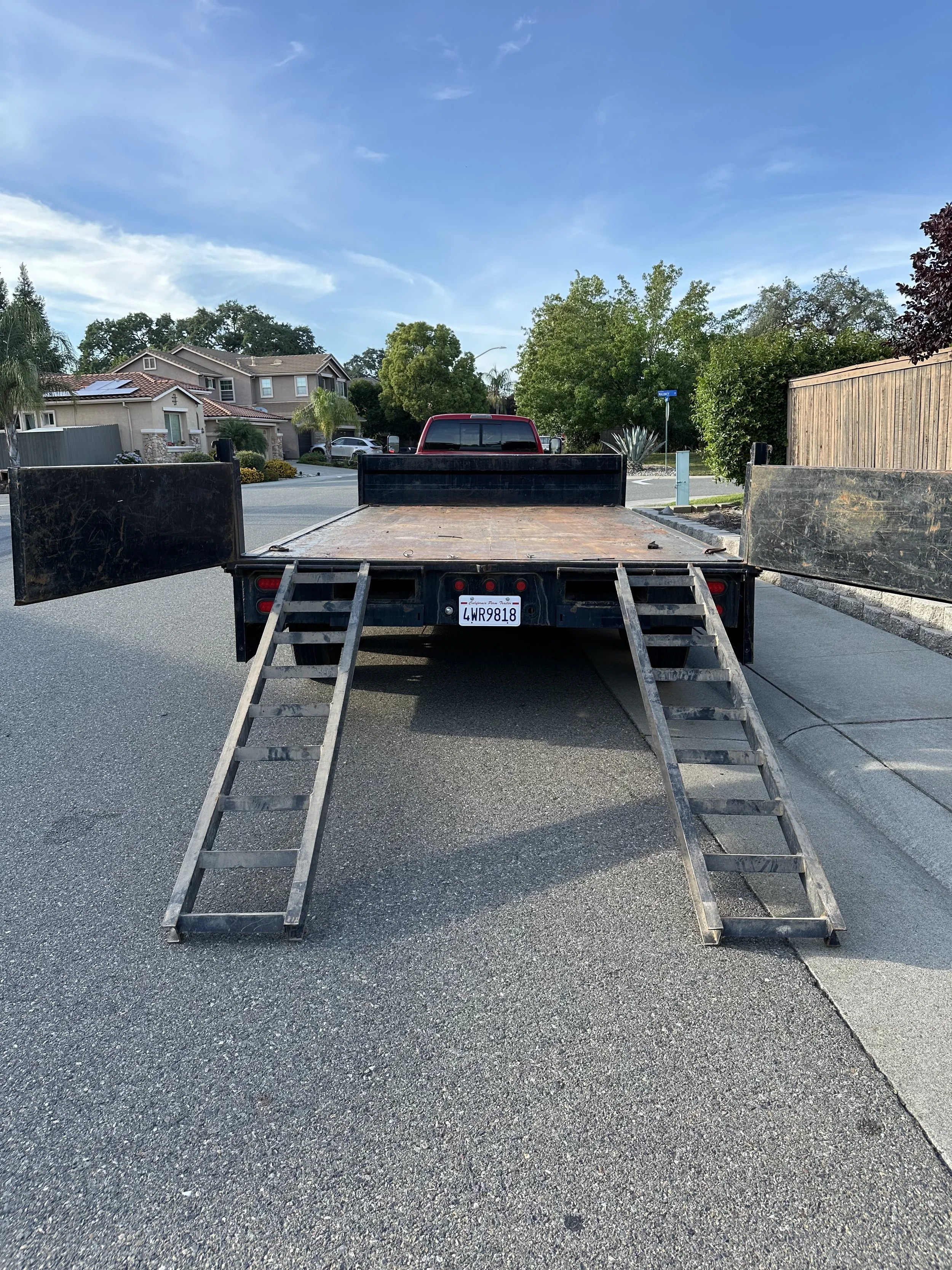 View of a truck with a flatbed and wooden ramps parked on a residential street, with houses, trees, and a blue sky in the background.