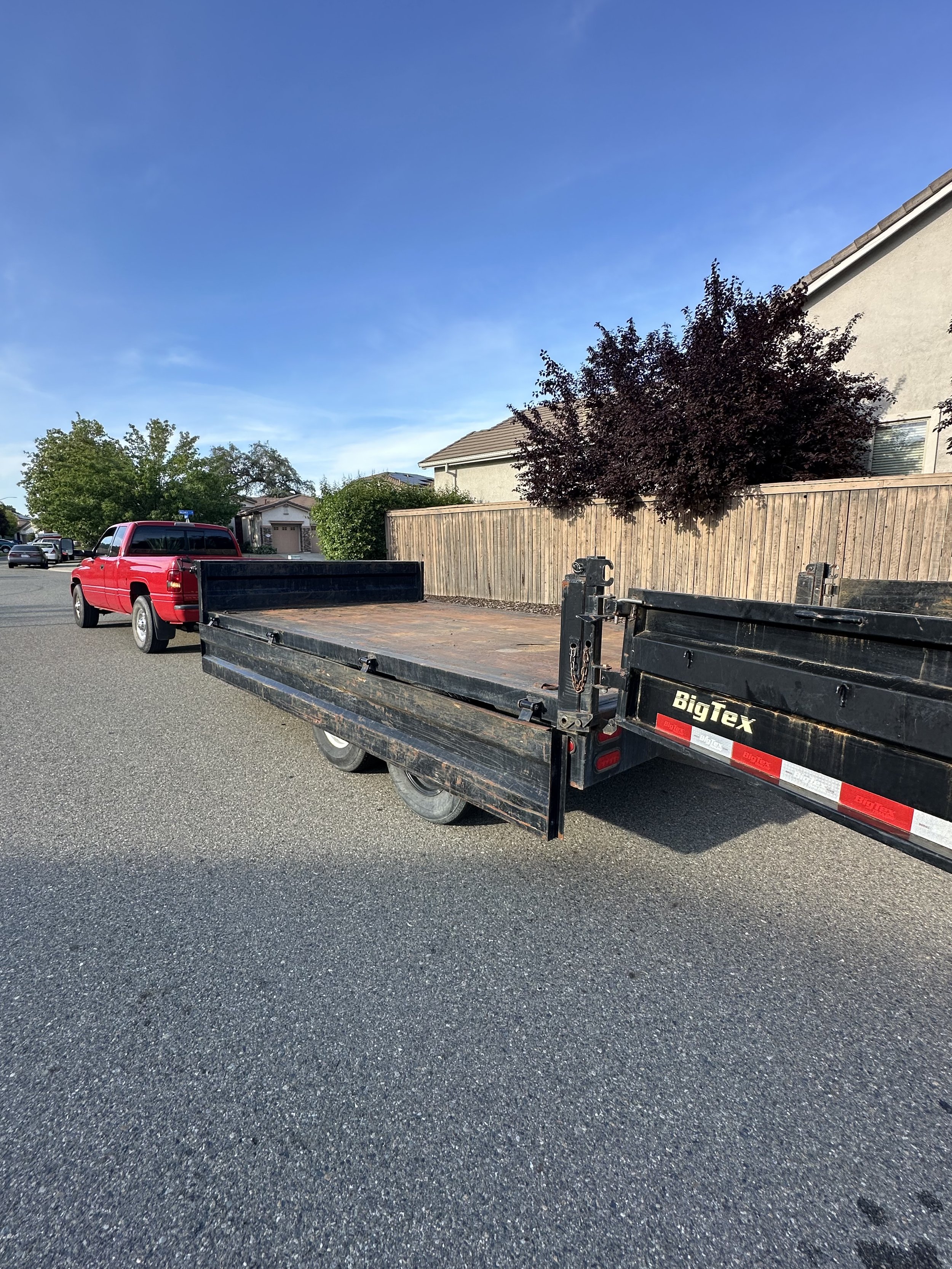 A black flatbed trailer attached to a red pickup truck parked on a residential street. Houses with trees and a clear blue sky in the background.