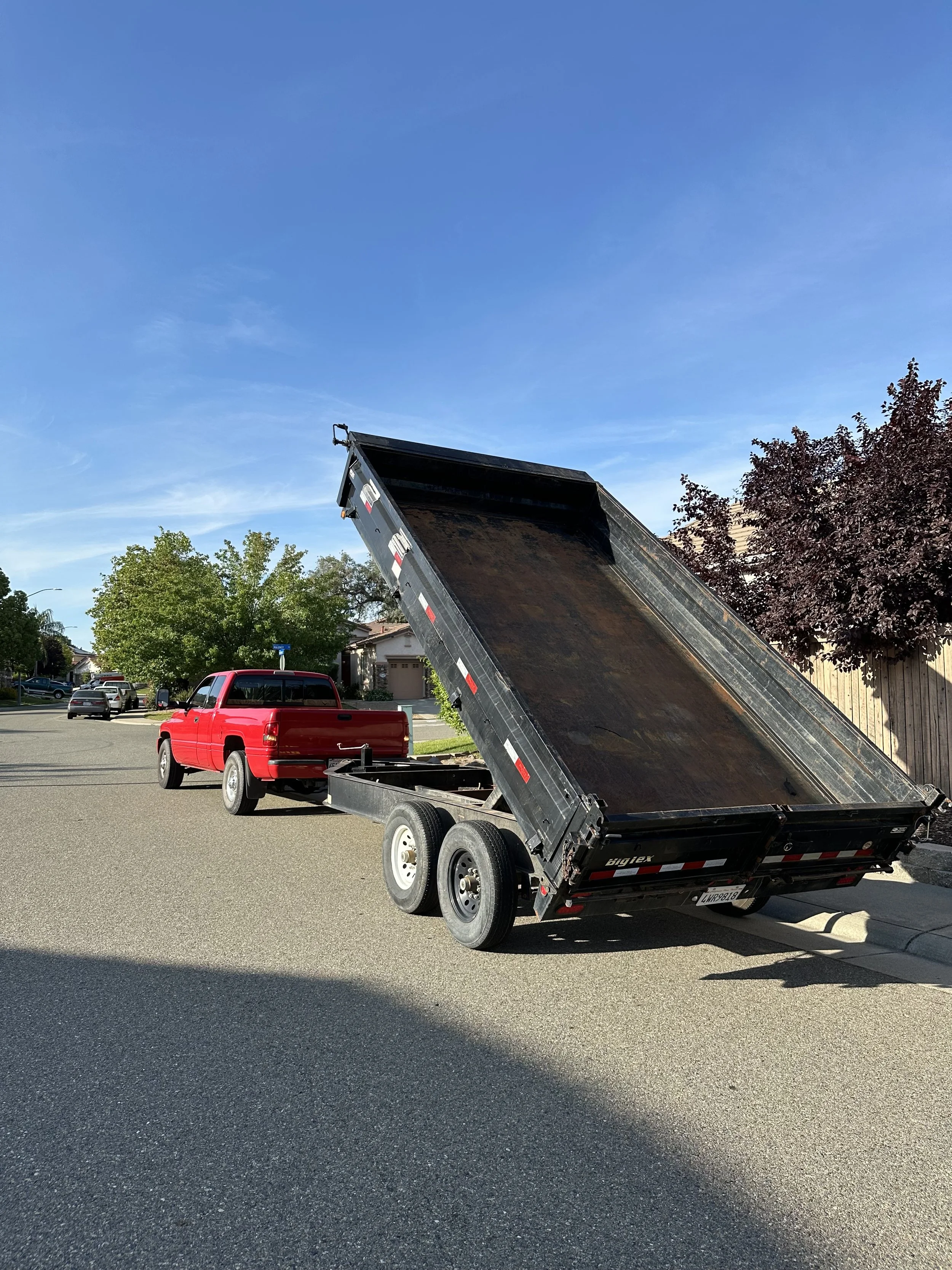 A red pickup truck parked on a residential street with a black dump trailer raised, showing its empty bed, under a clear blue sky with trees and houses in the background.