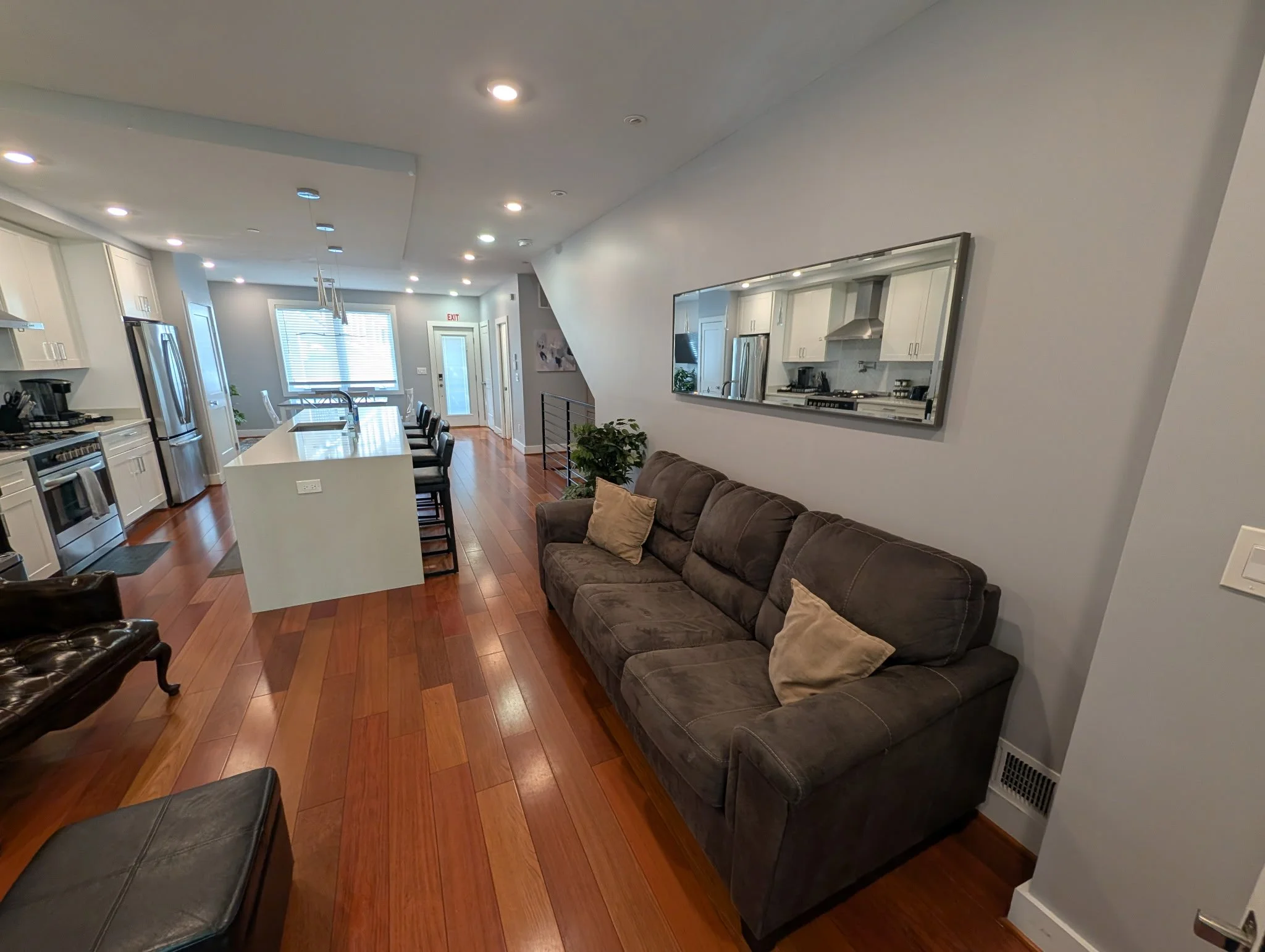 Interior of a modern kitchen and living room area with white cabinets, stainless steel appliances, a large kitchen island with bar stools, a brown sofa, and a large mirror on the wall.