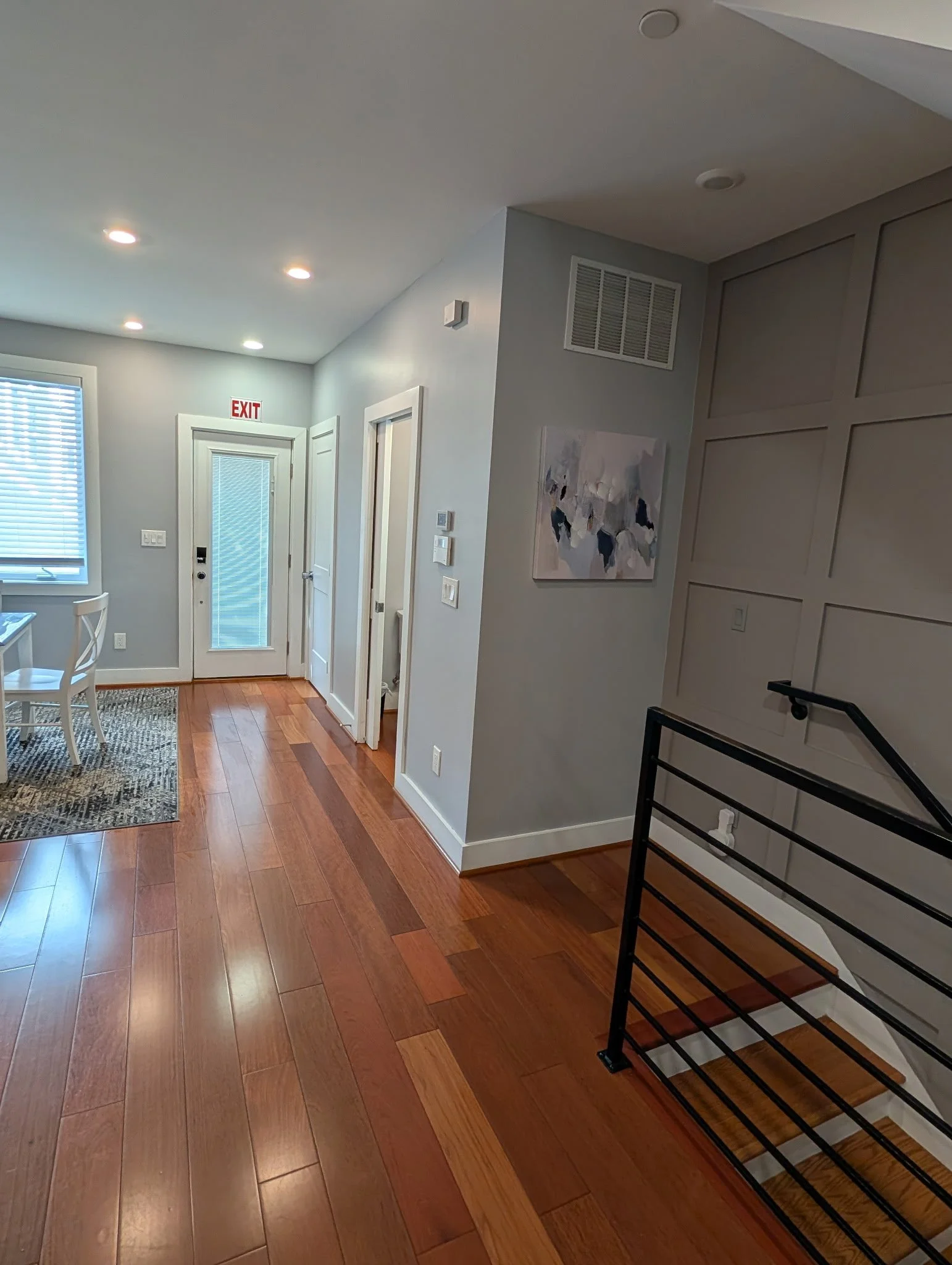 Interior view of a modern home entryway with hardwood flooring, white walls, a ceiling with recessed lighting, a staircase with a metal railing, a framed abstract artwork on the wall, a window with blinds, and an exit door with glass panels.