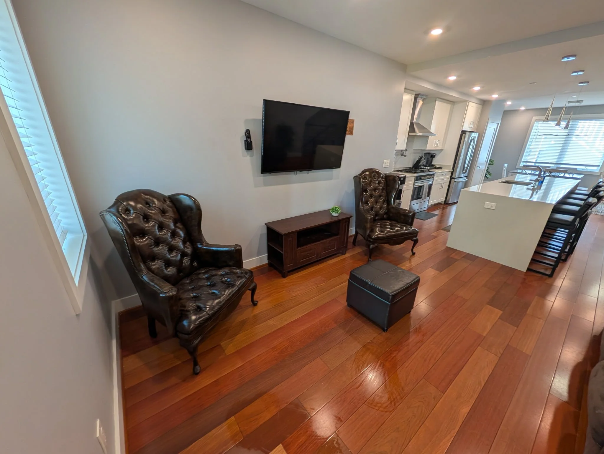 Living room and kitchen area with hardwood floors, two leather armchairs, a wall-mounted flat-screen TV, a small side table with a plant, kitchen appliances including a refrigerator, stove, and dishwasher, a kitchen island with barstools, and bright natural light from windows.