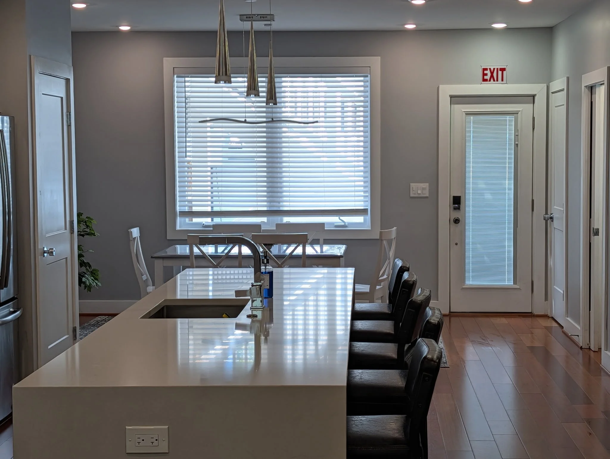 A modern kitchen and dining area with a white island countertop, black chairs, a large window with white blinds, and a door with blinds. There are pendant lights hanging from the ceiling and a small potted plant on the left side. The room has gray walls and wooden flooring.