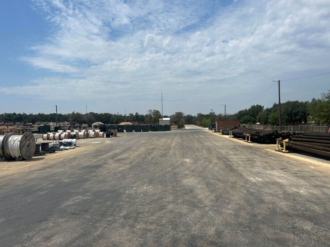 An outdoor industrial yard with metal pipes and spools laid out on both sides of a paved road under a partly cloudy sky.