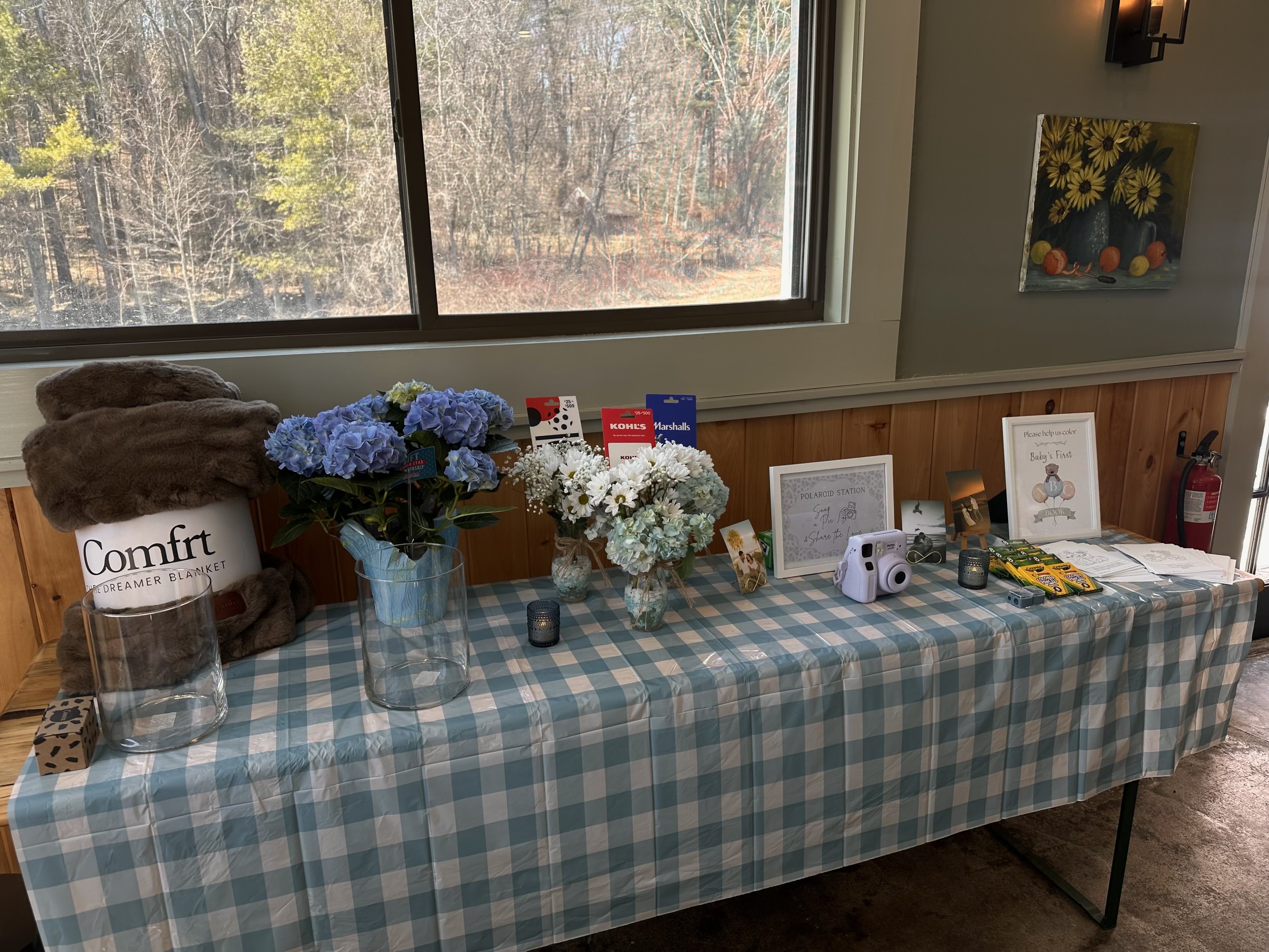 A table covered with a blue and white checkered tablecloth displaying various floral arrangements, candles, and framed signs near a window overlooking a wooded area.