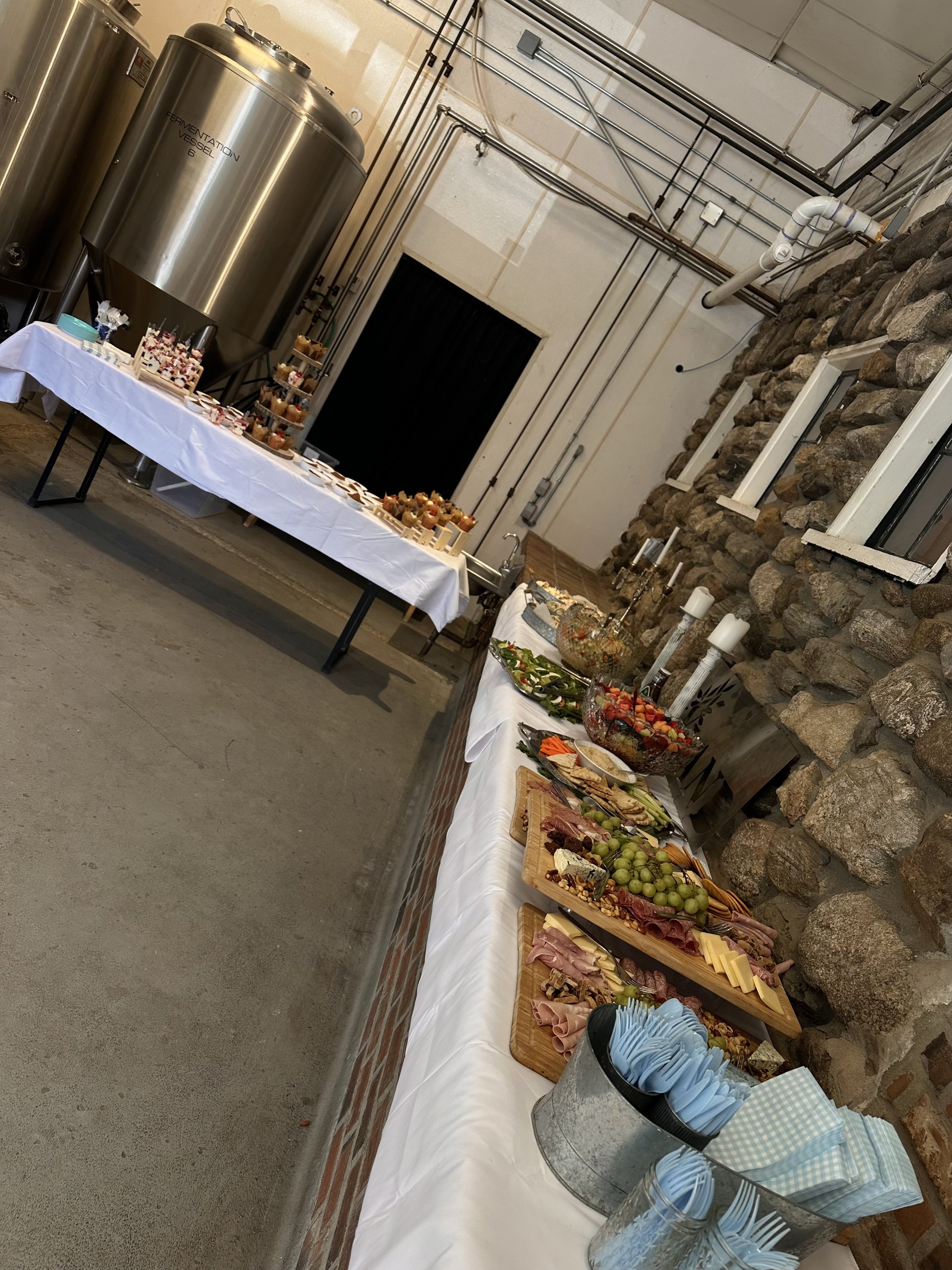 Buffet table with snacks and appetizers including cheeses, meats, fruits, vegetables, and finger foods, set against a stone wall in a rustic setting. Large metal fermentation tanks are visible in the background.